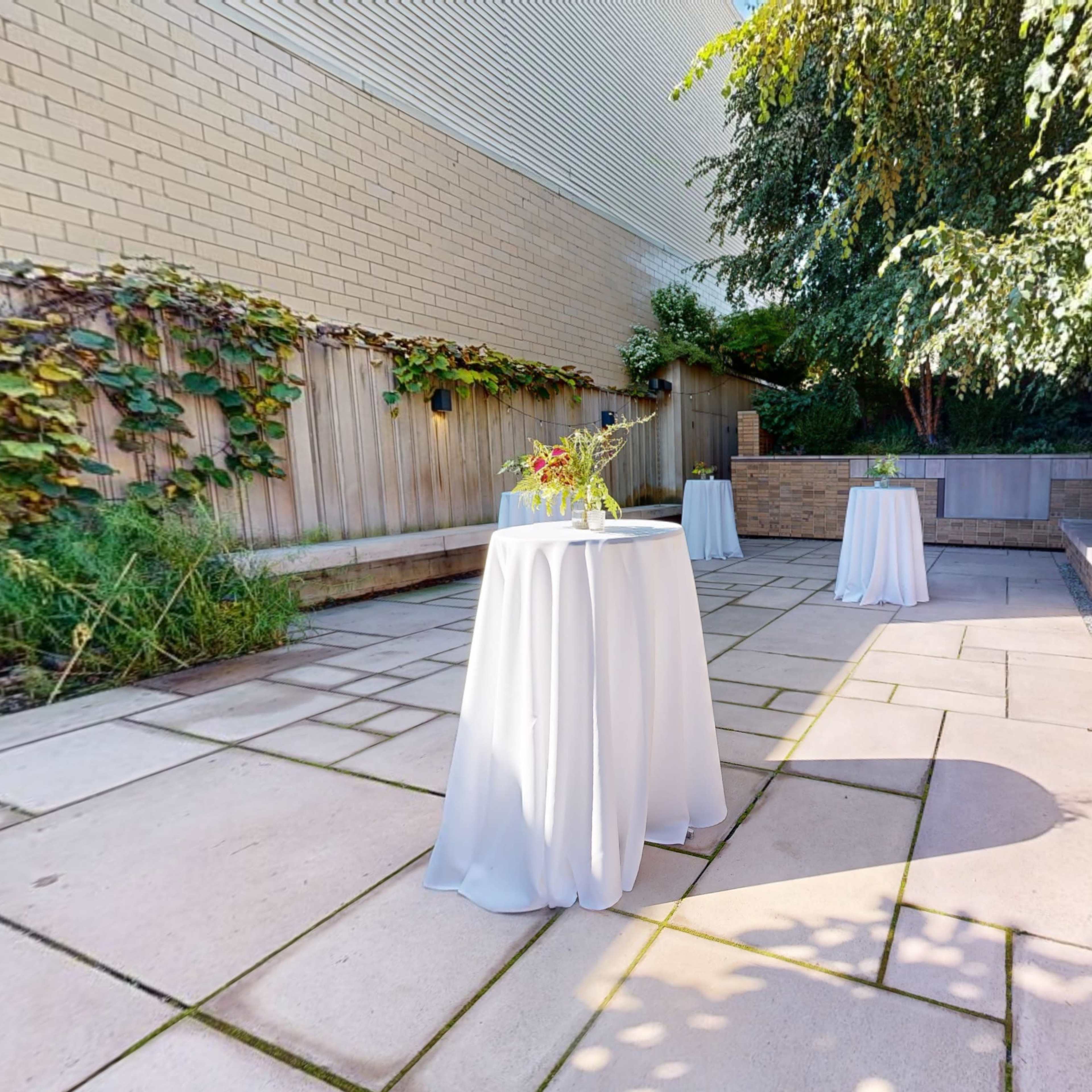 A courtyard with three round tables covered in white tablecloths, surrounded by greenery and stone pathways.