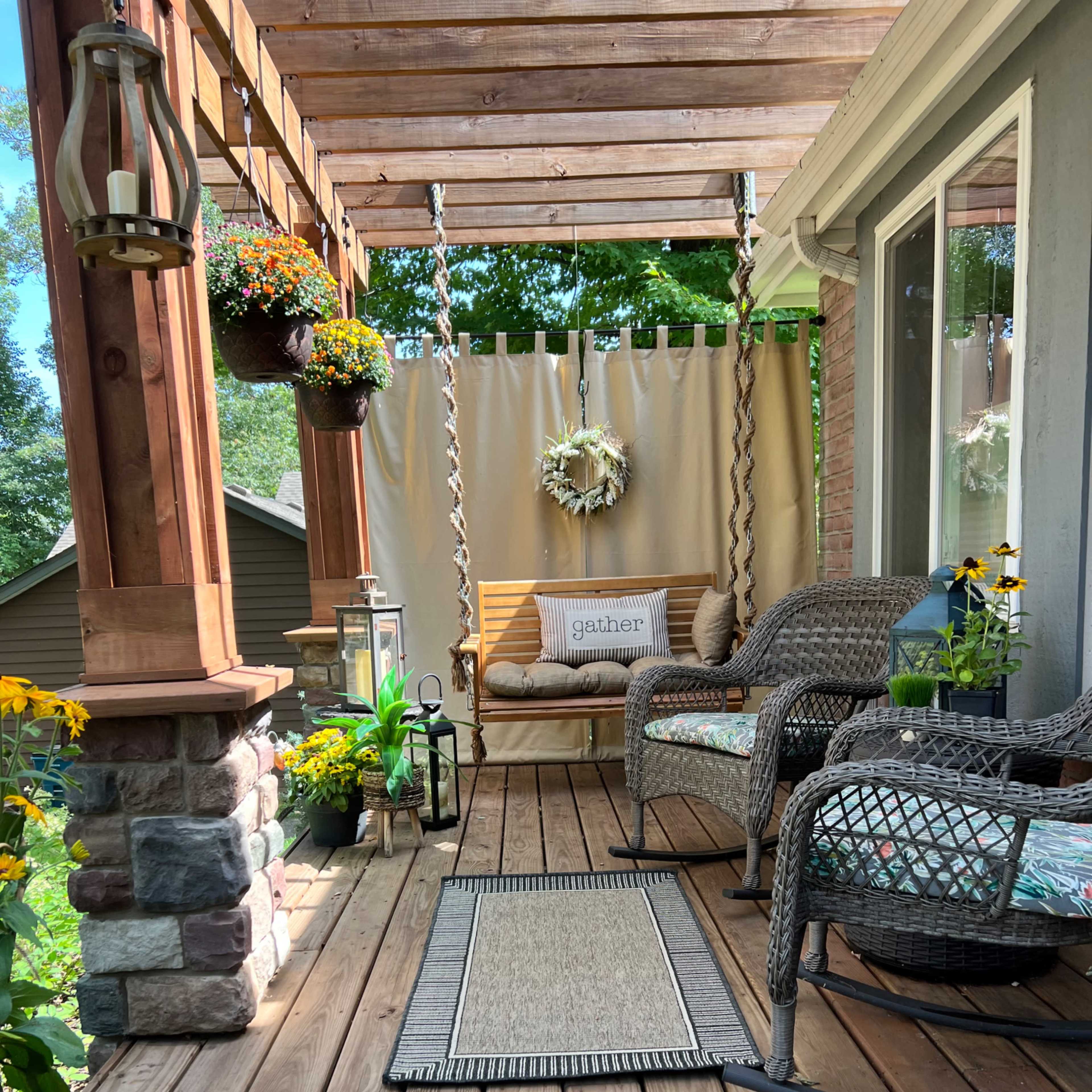 A porch with wooden flooring, two wicker chairs, potted plants, and a swing draped with a curtain.