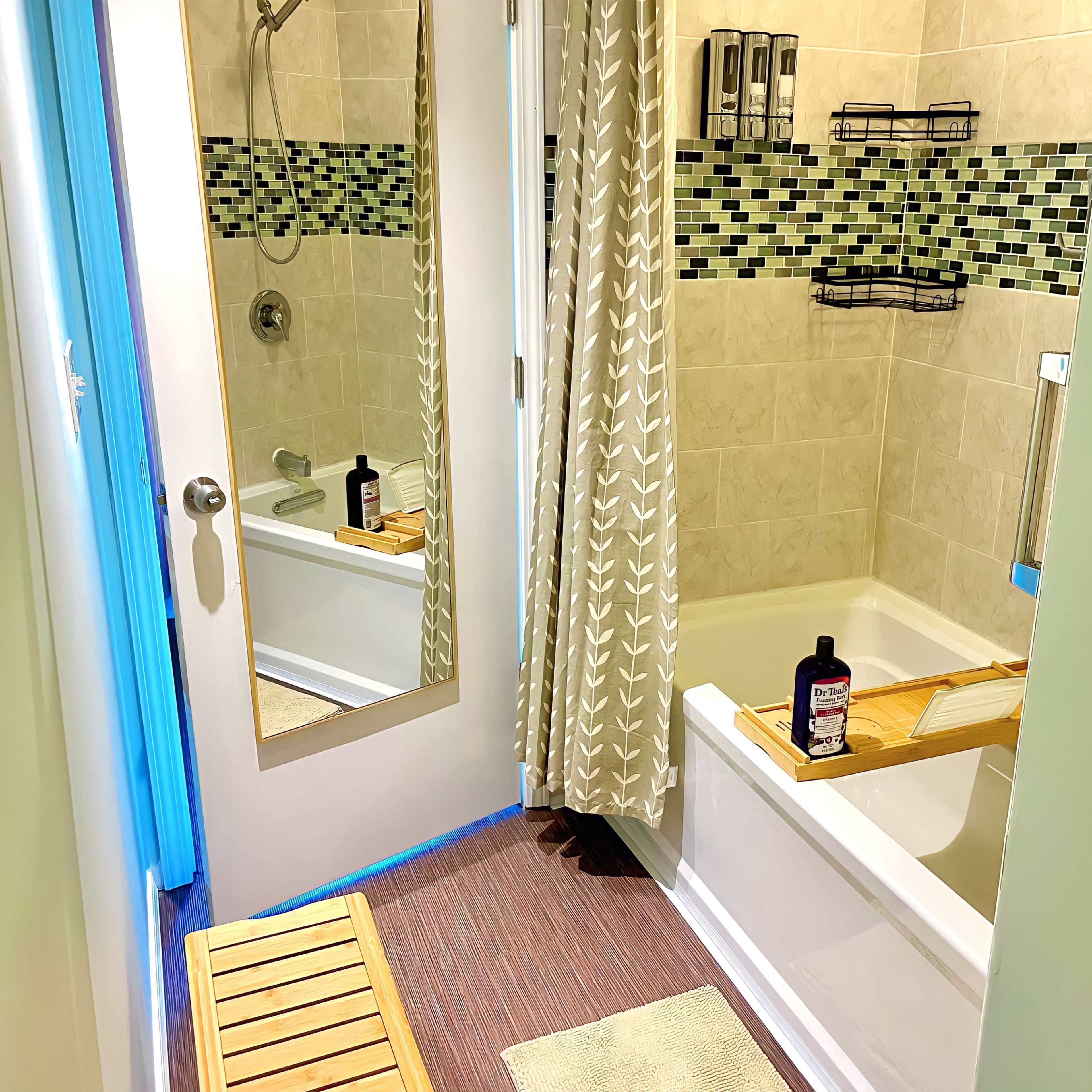 A bathroom features a bathtub with a wooden bath tray, a shower with glass doors, and a mirror reflecting a doorway.