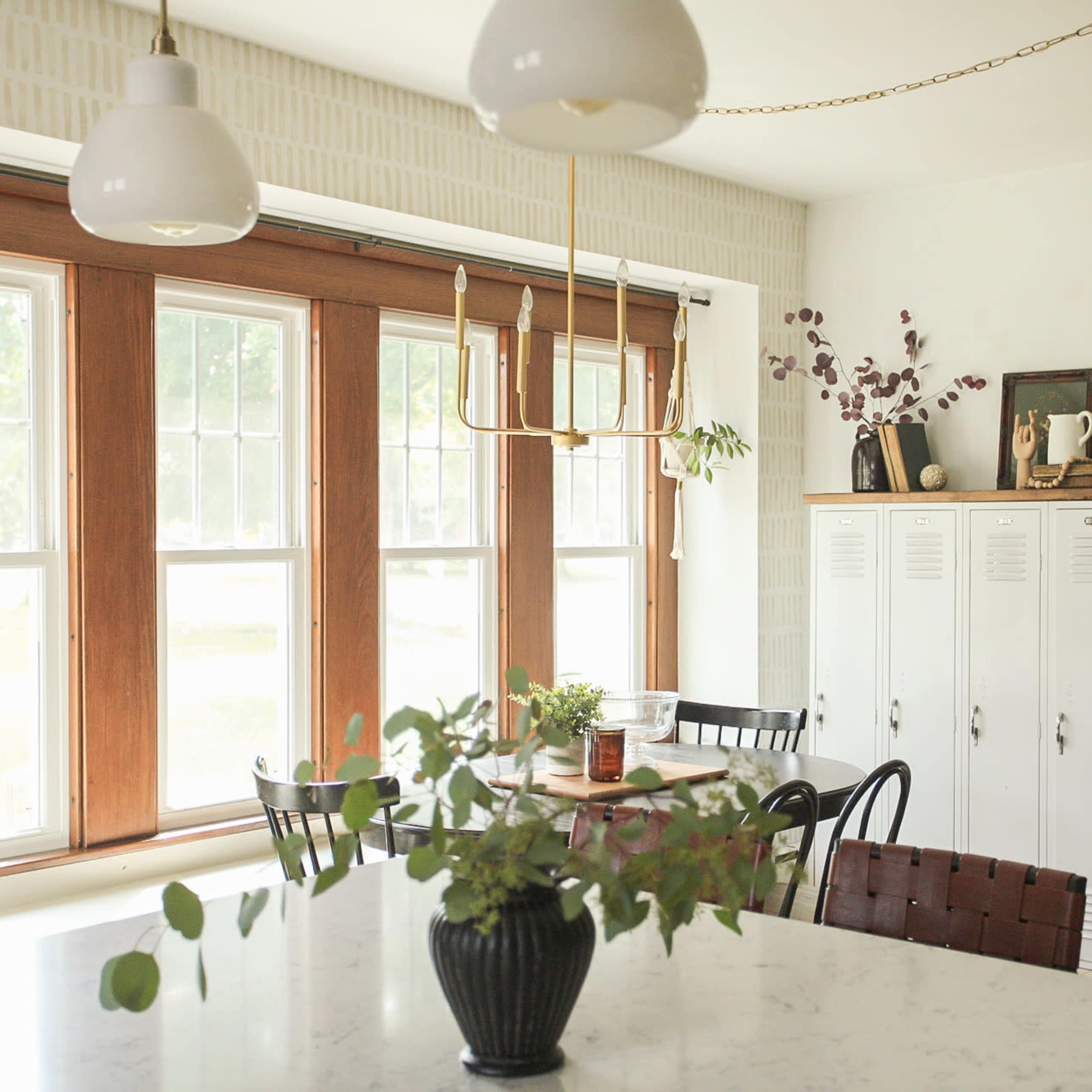 A bright kitchen features large windows, a dining table, black chairs, a white cabinet, and a vase with greenery on the counter.
