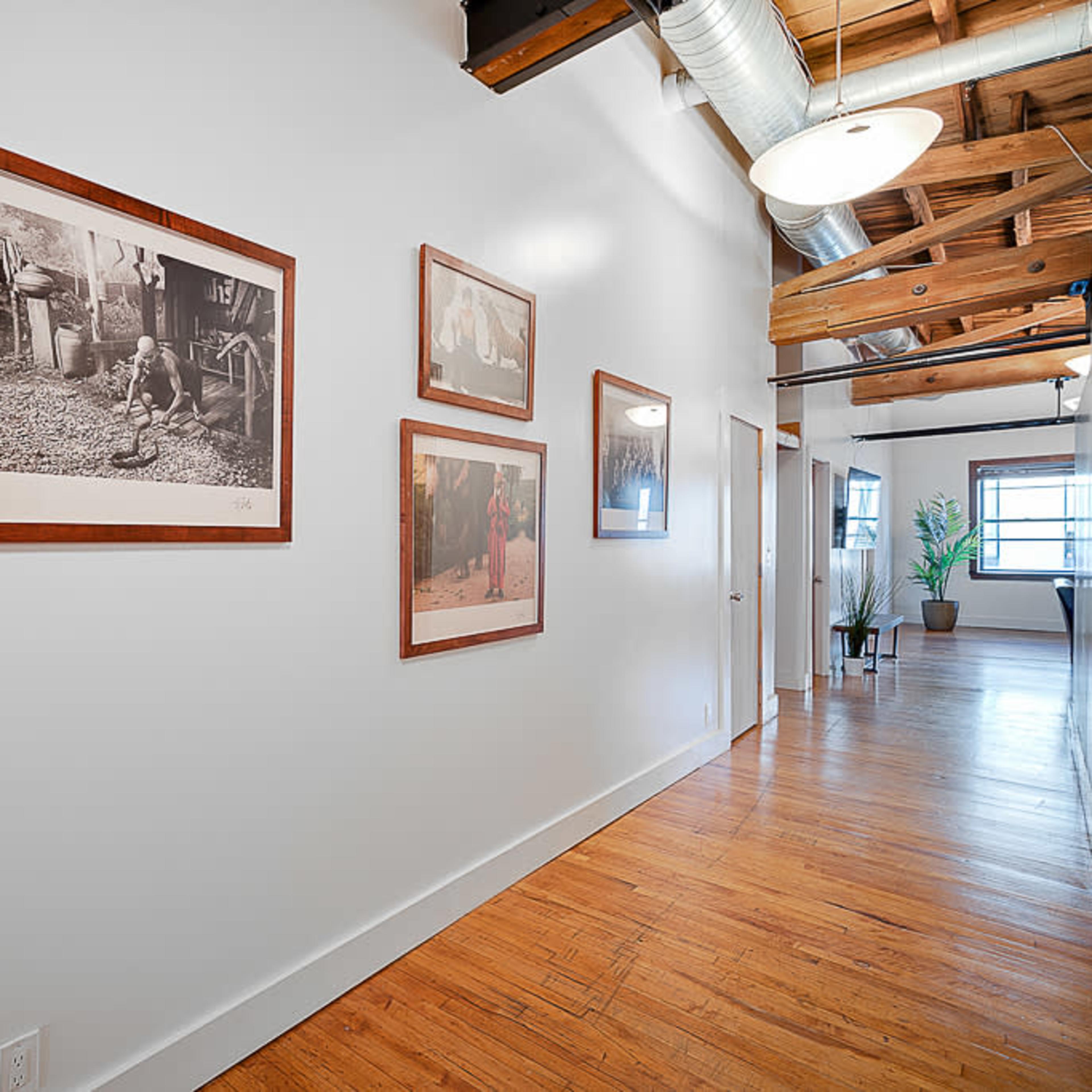 A hallway with framed photographs on the wall and wooden flooring, featuring exposed beams and overhead lighting.