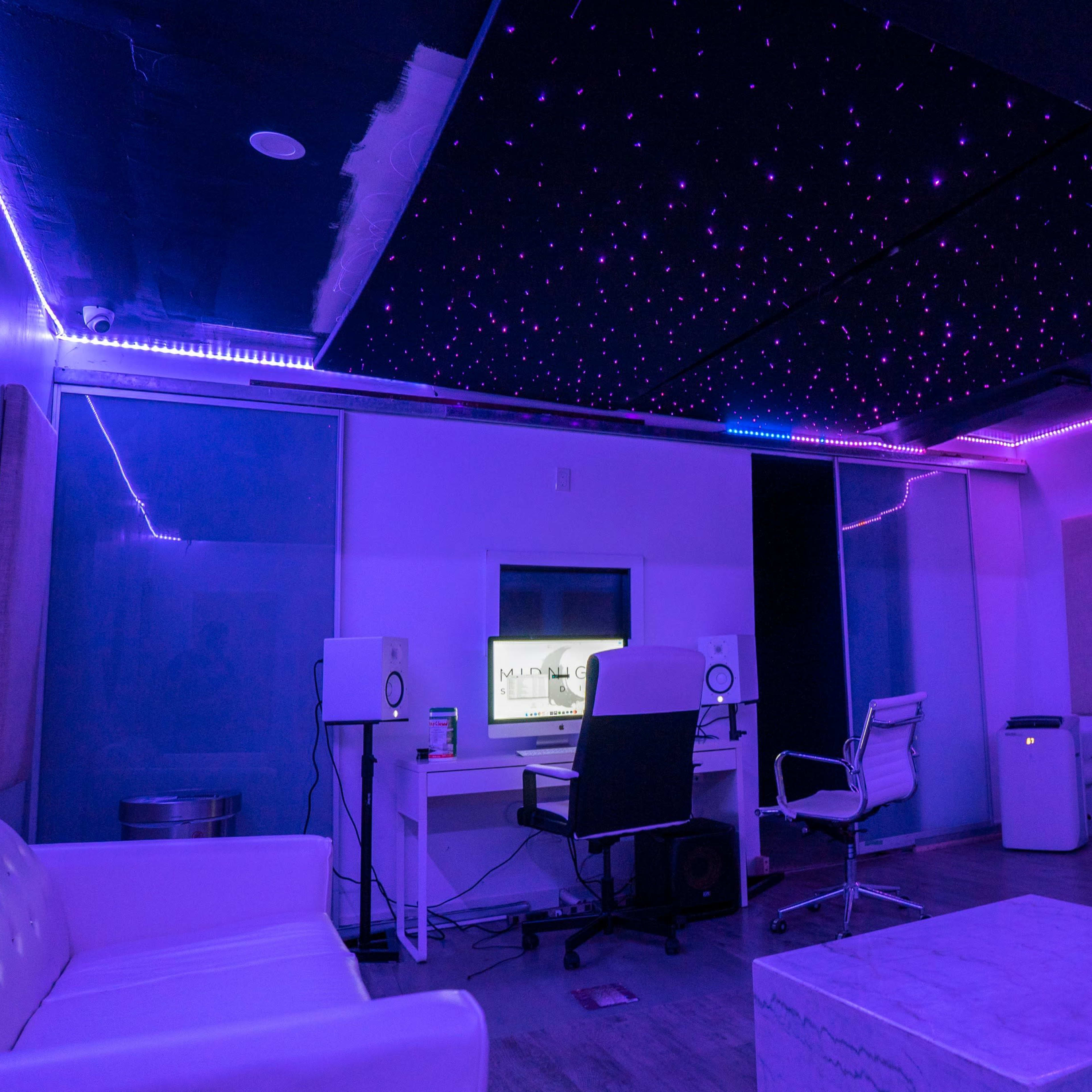 A modern recording studio with purple lighting, a white sofa, a desk with studio monitors, and a screen in the background.