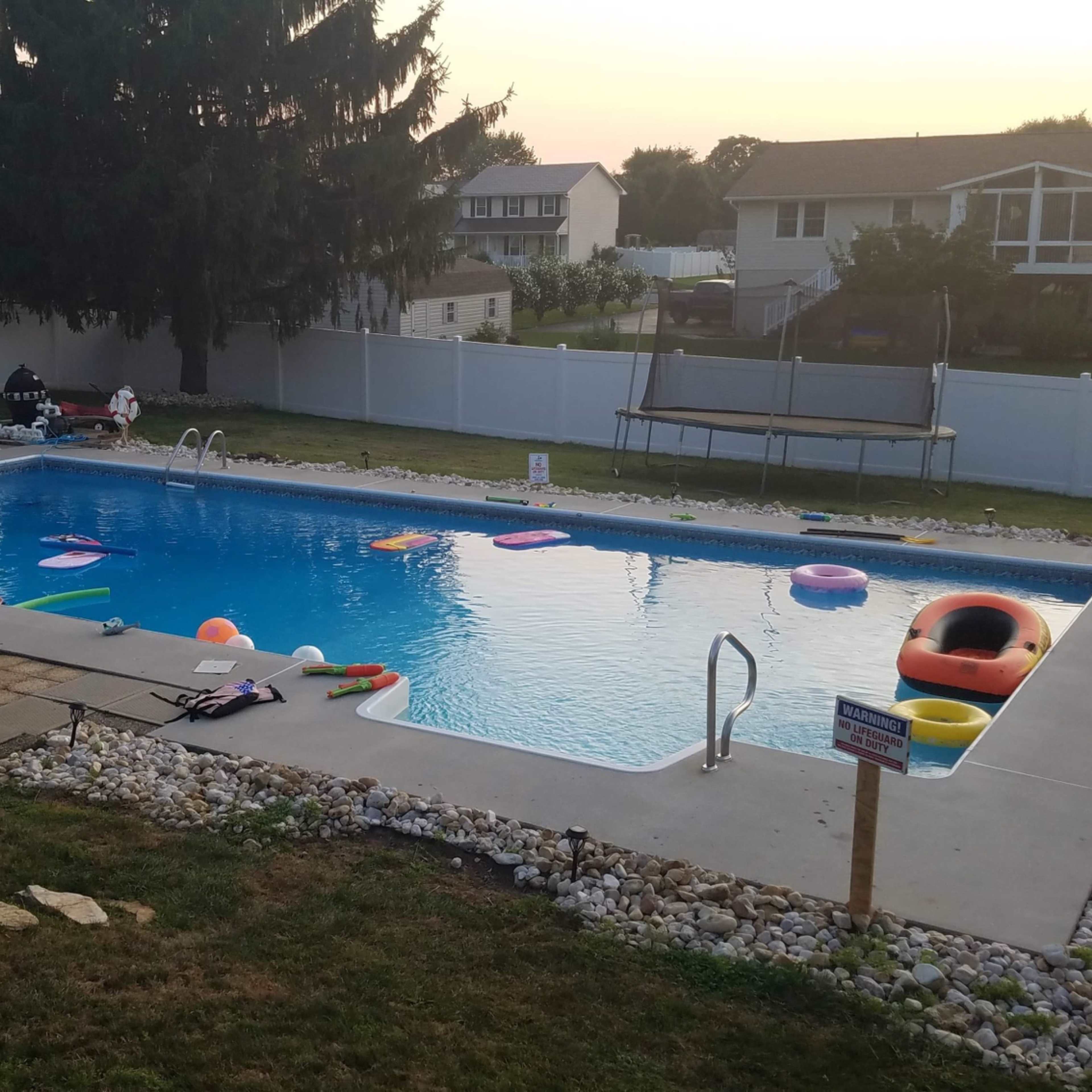 A backyard swimming pool surrounded by colorful inflatable floaties, a wooden deck, and a trampoline in a fenced yard at sunset.
