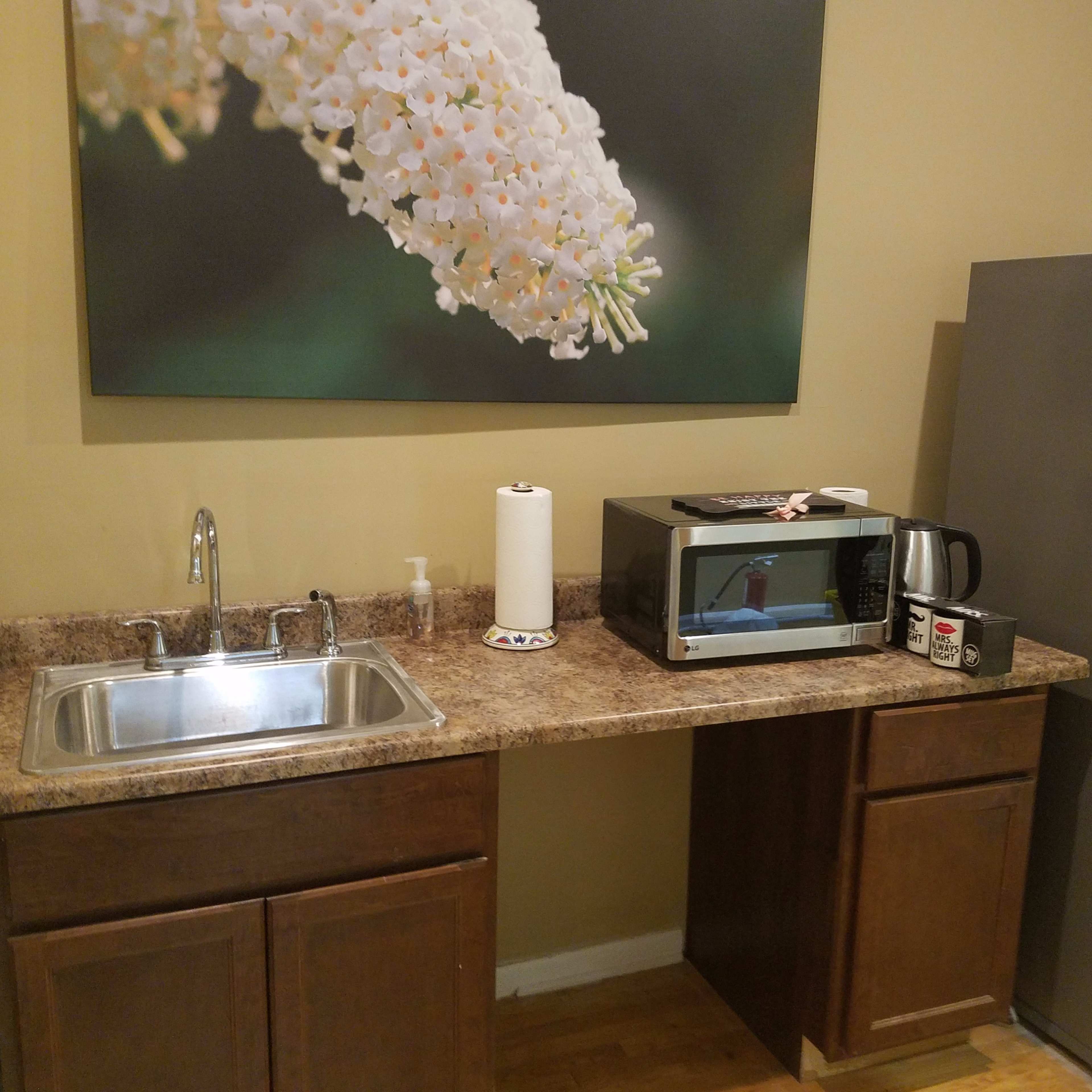 A kitchen area with a sink, a microwave, a kettle, and a silver refrigerator against a beige wall with a large floral photograph.