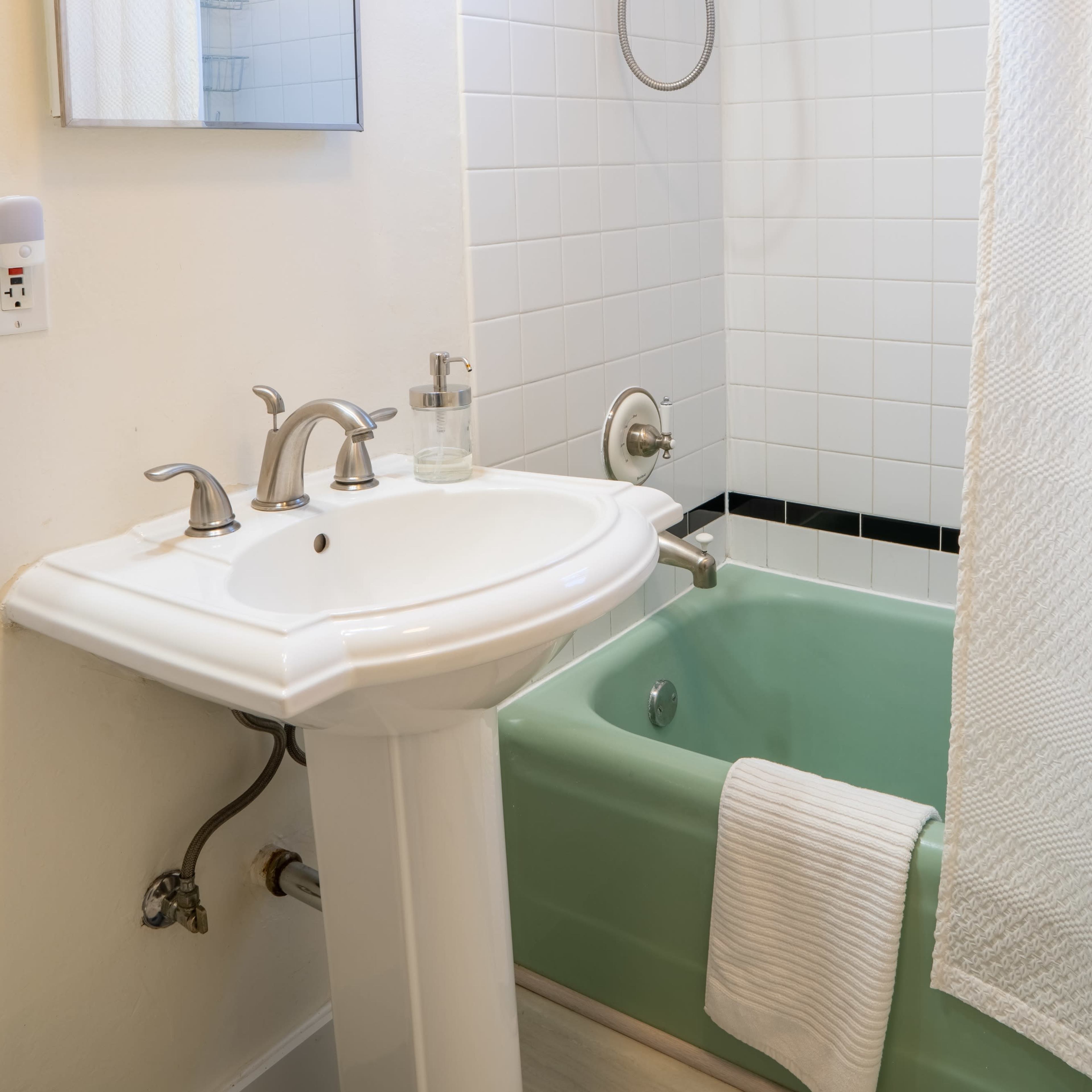 A bathroom with a white tiled wall, a green bathtub, a pedestal sink, and a showerhead above the tub.