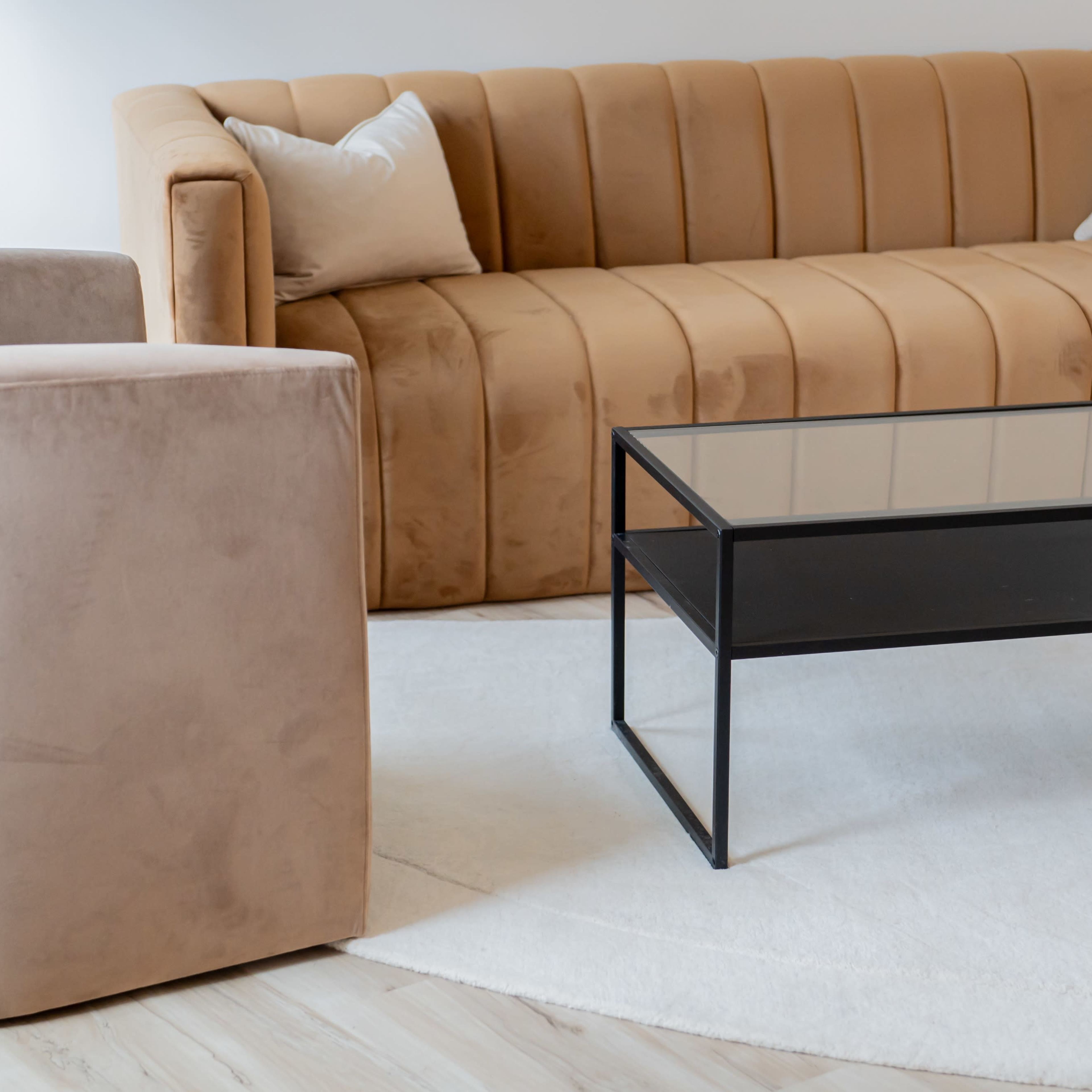 A modern living room with a tan velvet sofa, a beige armchair, and a black metal and glass coffee table on a white rug.