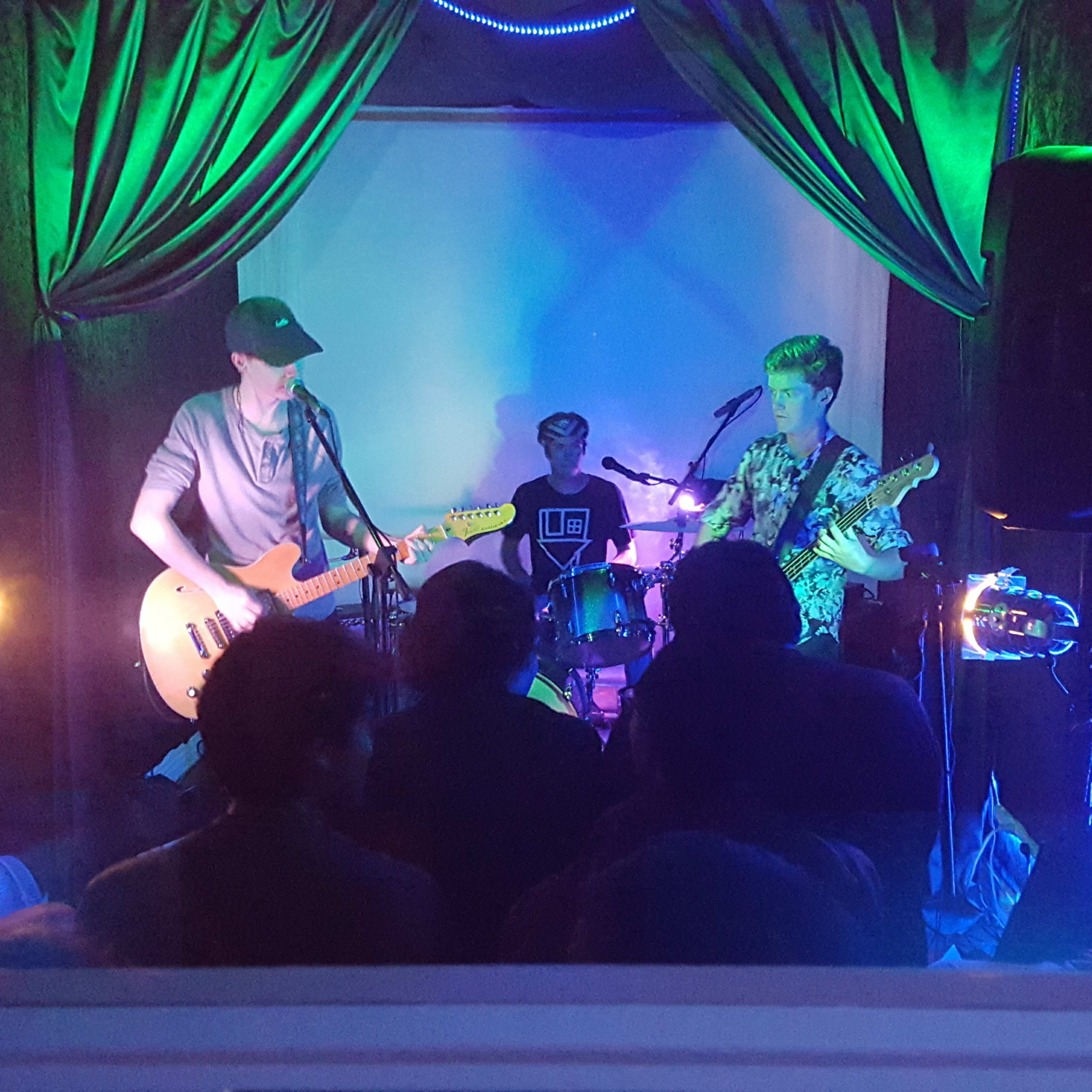 Two musicians play guitar and drums on a small stage with colorful lighting in front of an audience.