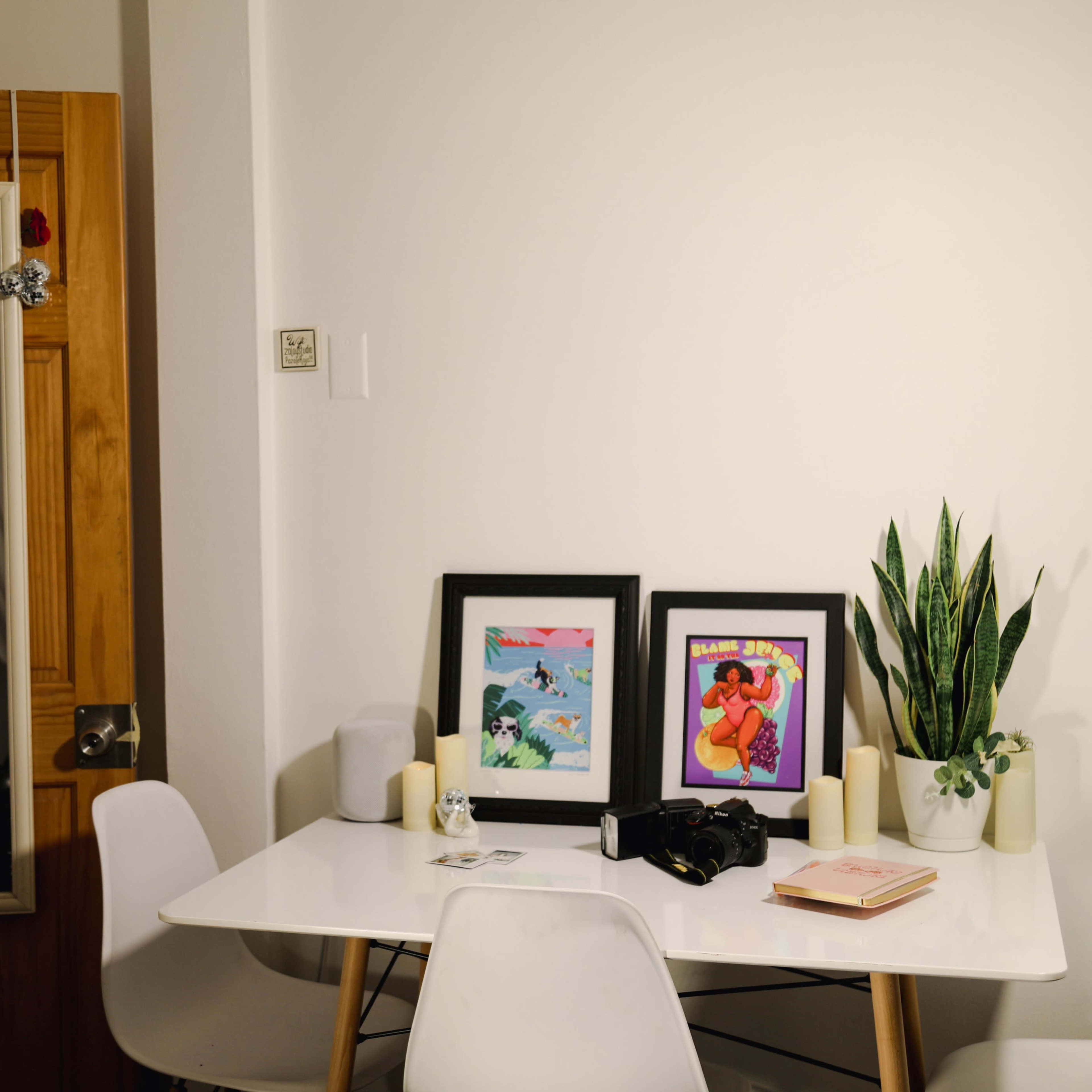 A white table is set up in a room with two framed pictures, candles, a plant, and a camera, while a mirror hangs on the wall beside a wooden door.