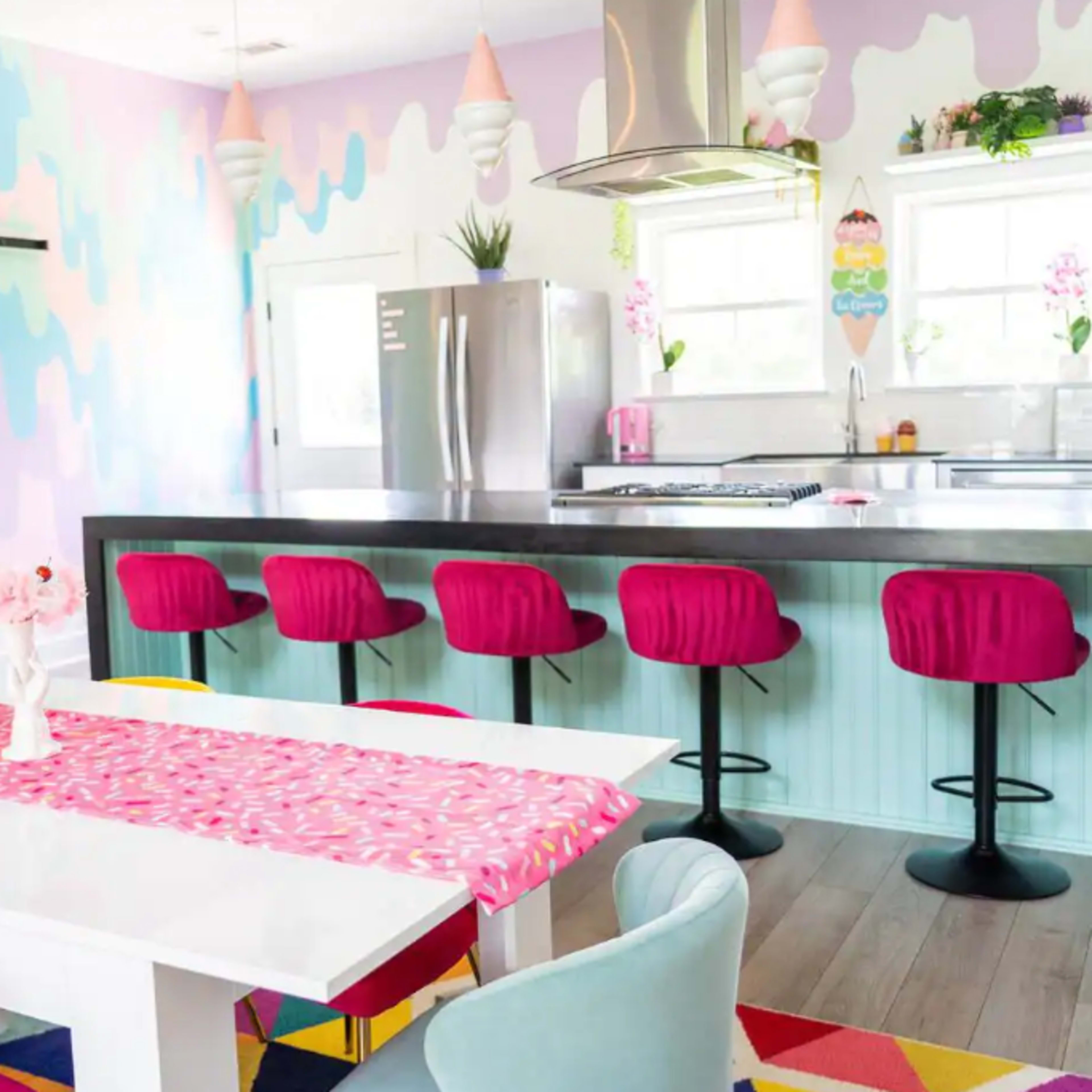 A brightly colored kitchen and dining area with a pastel-themed wall, modern stools at the counter, and a white dining table topped with a vibrant table runner.