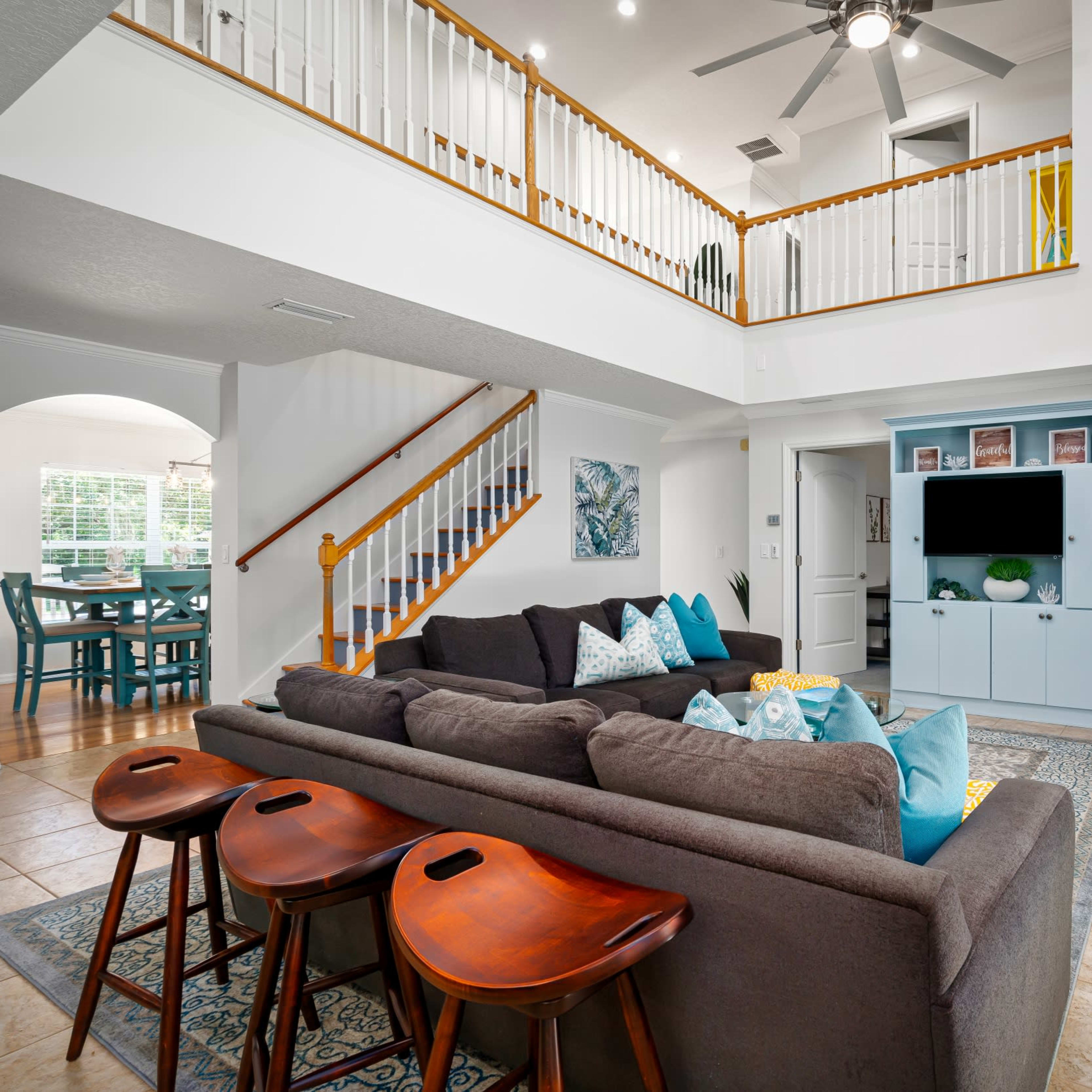 A spacious living room features a gray sectional sofa with colorful pillows, a wooden coffee table, and an entertainment unit, overlooking a dining area with a staircase in the background.