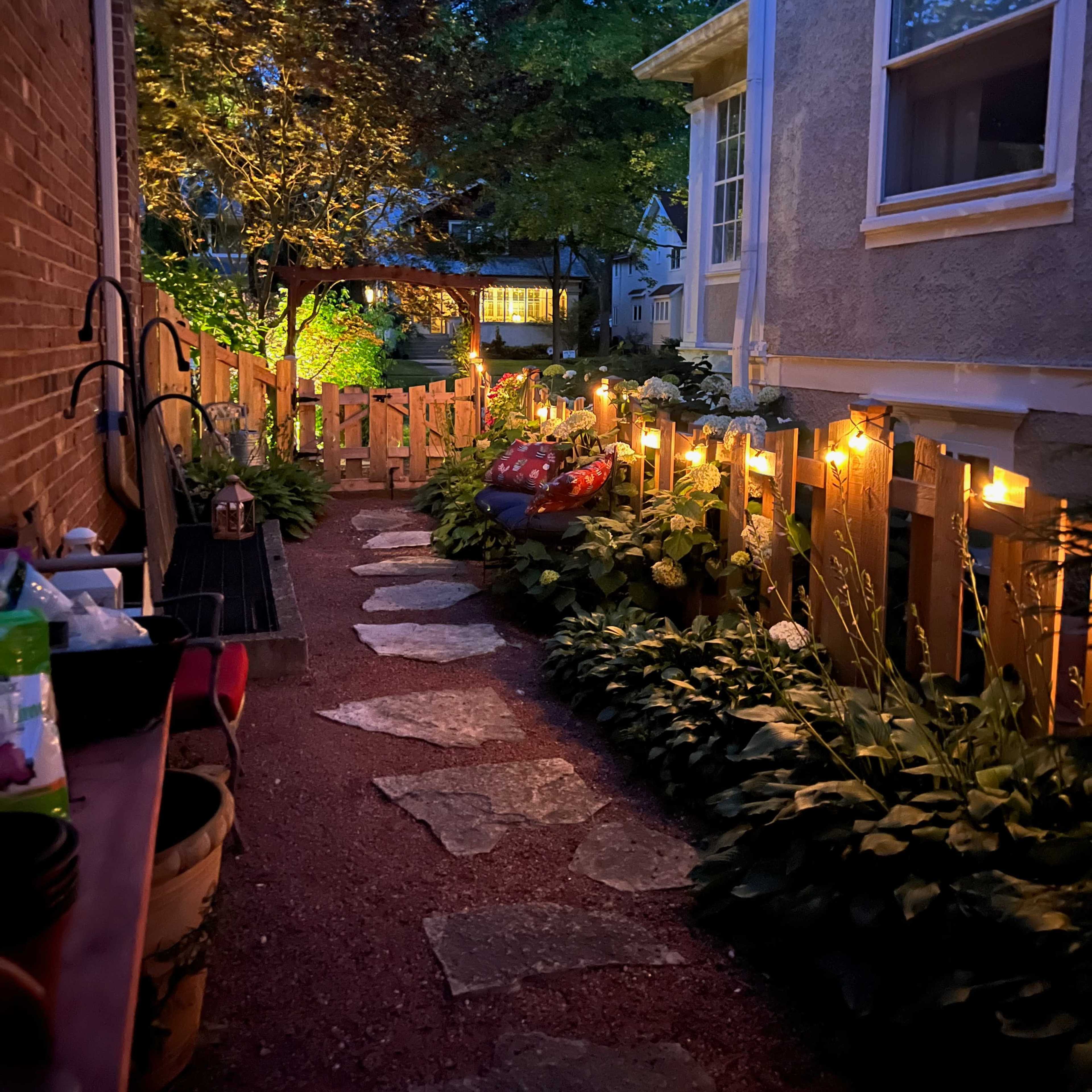 A stone path lined with greenery and illuminated by string lights runs alongside a house in the evening.