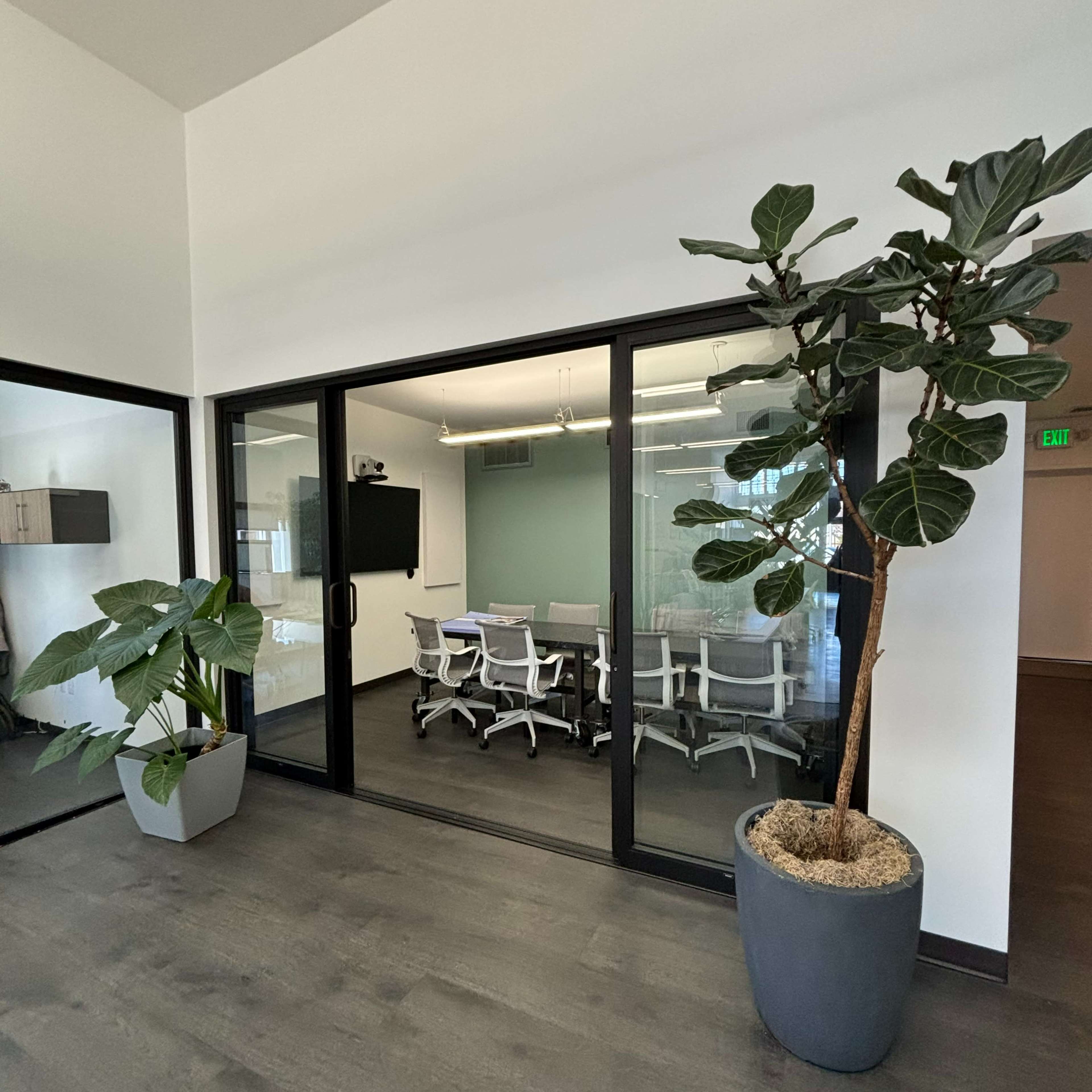 A modern conference room is visible through glass walls, featuring a large table surrounded by white chairs and decorated with potted plants outside.