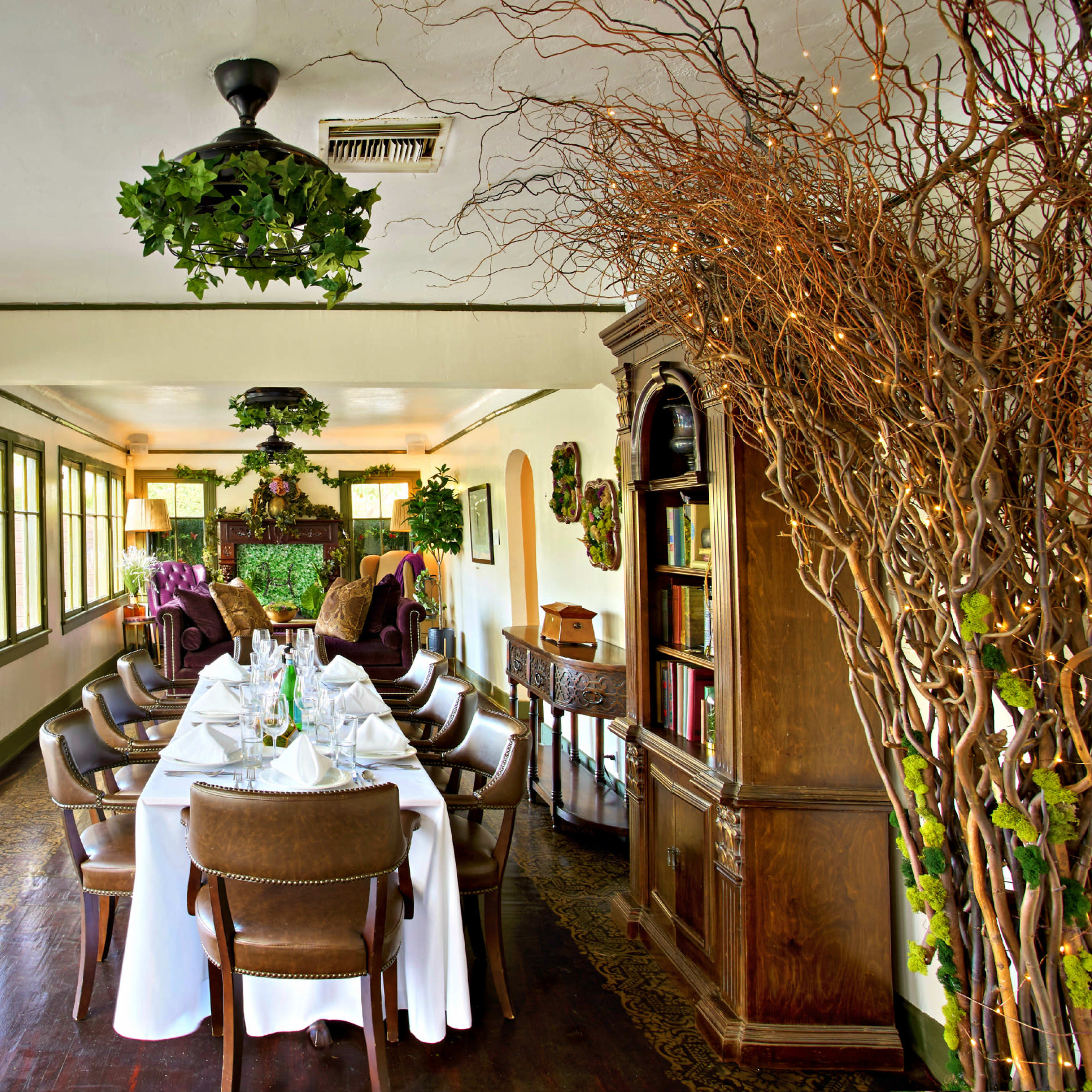 A long dining table is set with white tablecloths in a quaint room adorned with plants and wooden furniture.