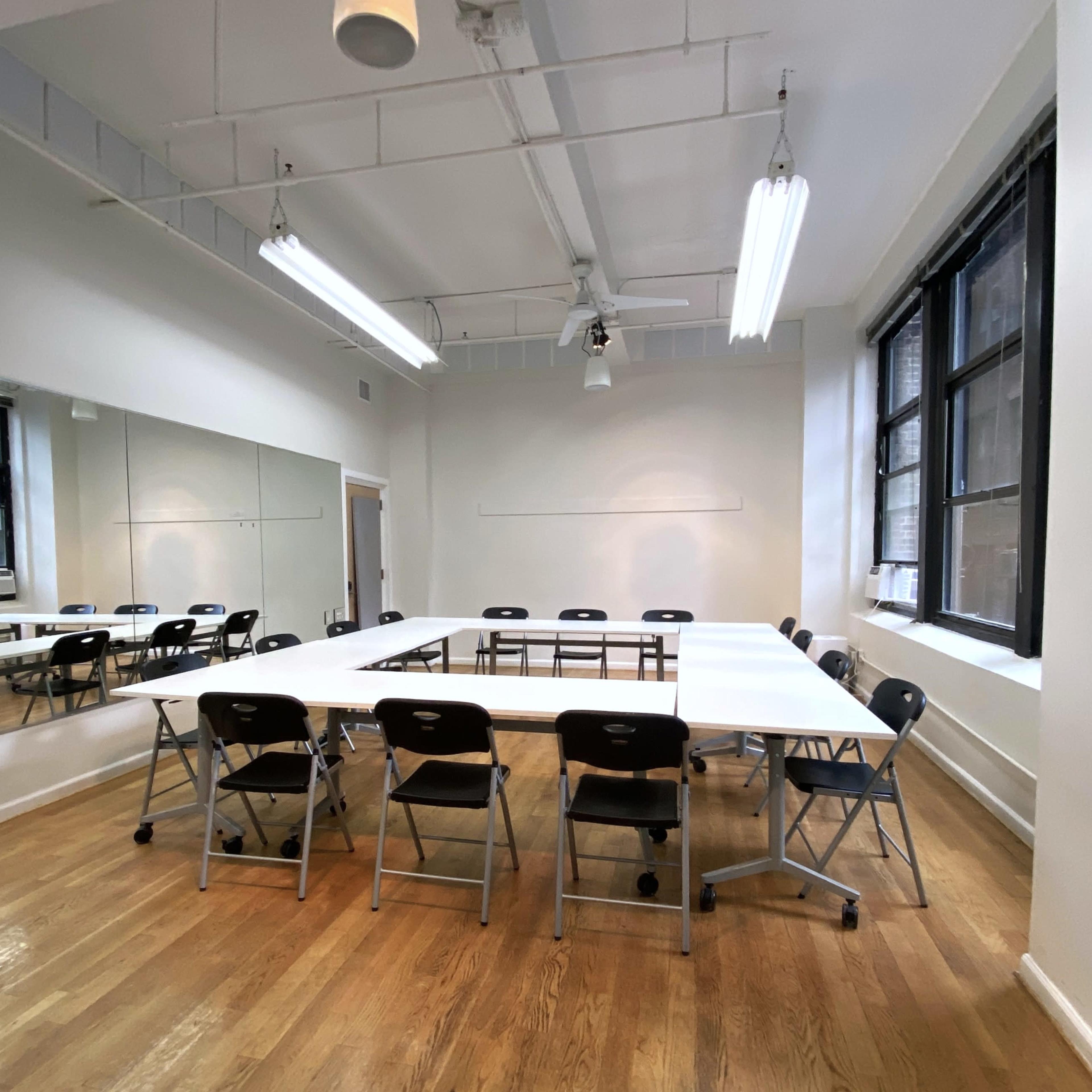 A bright, minimalistic meeting room features a large rectangular table surrounded by folding chairs, with windows and mirrors lining the walls.
