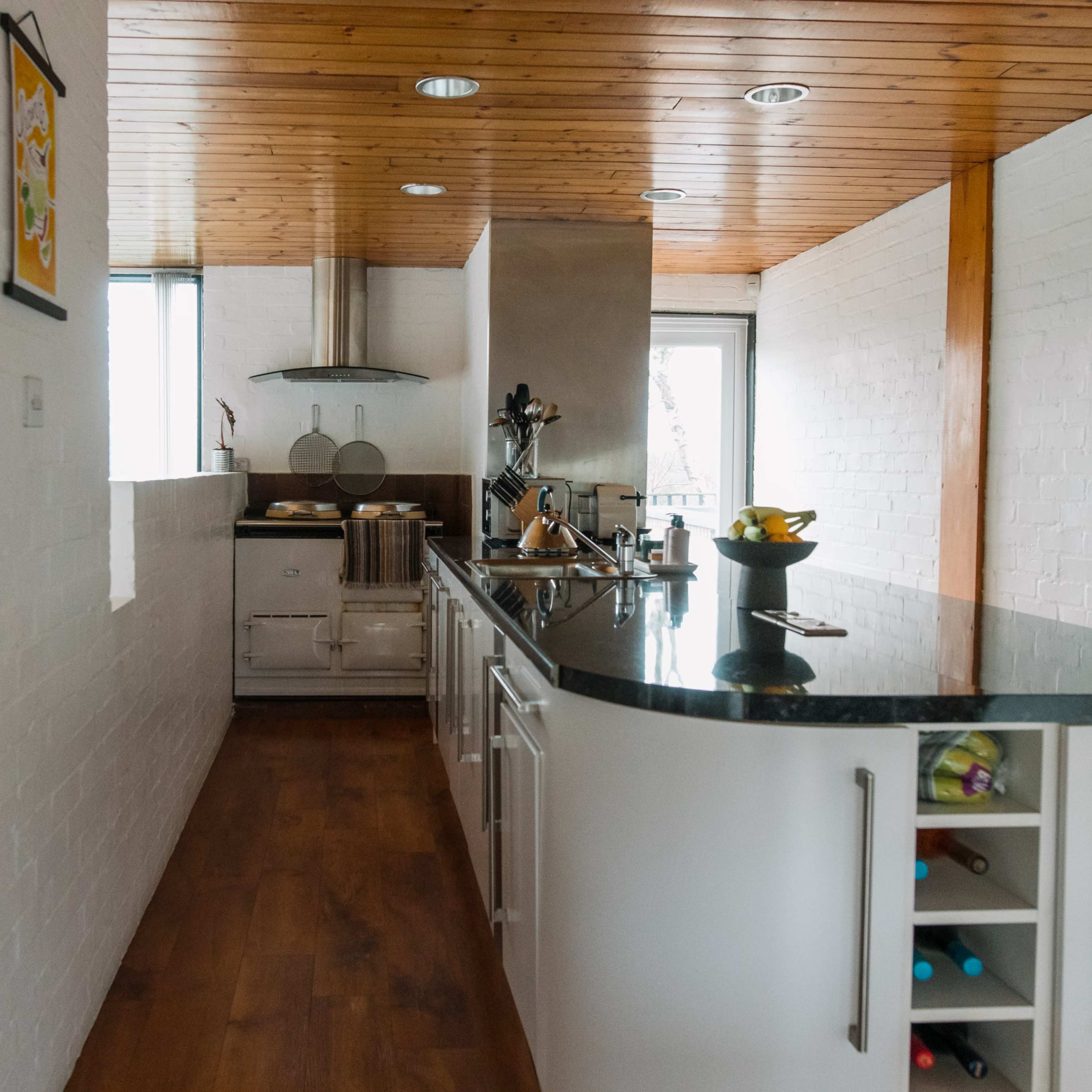 A modern kitchen features a long countertop, wooden ceiling, and large windows providing natural light.