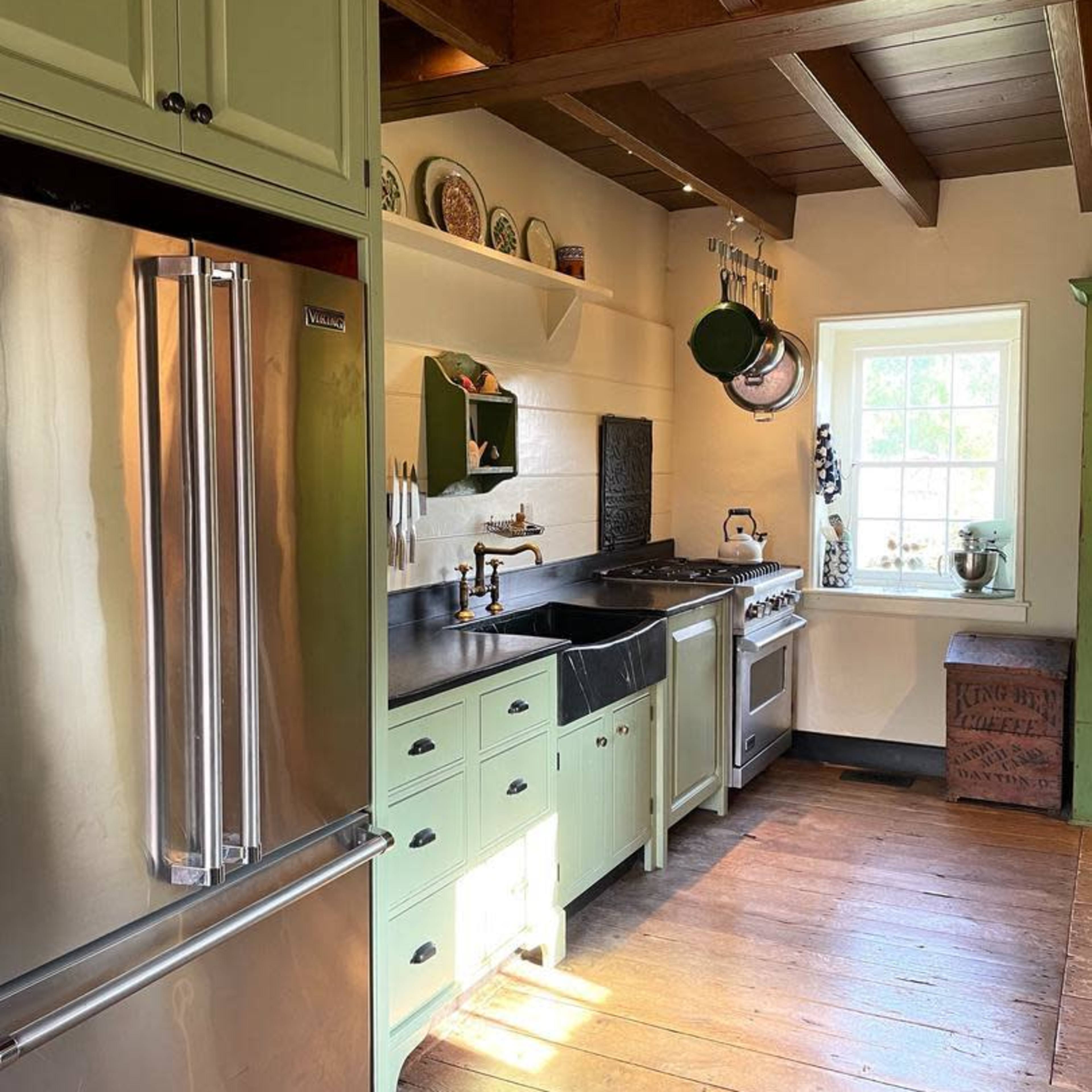 The kitchen features green cabinets, stainless steel appliances, a black countertop, and wooden beams overhead, illuminated by natural light from a window.