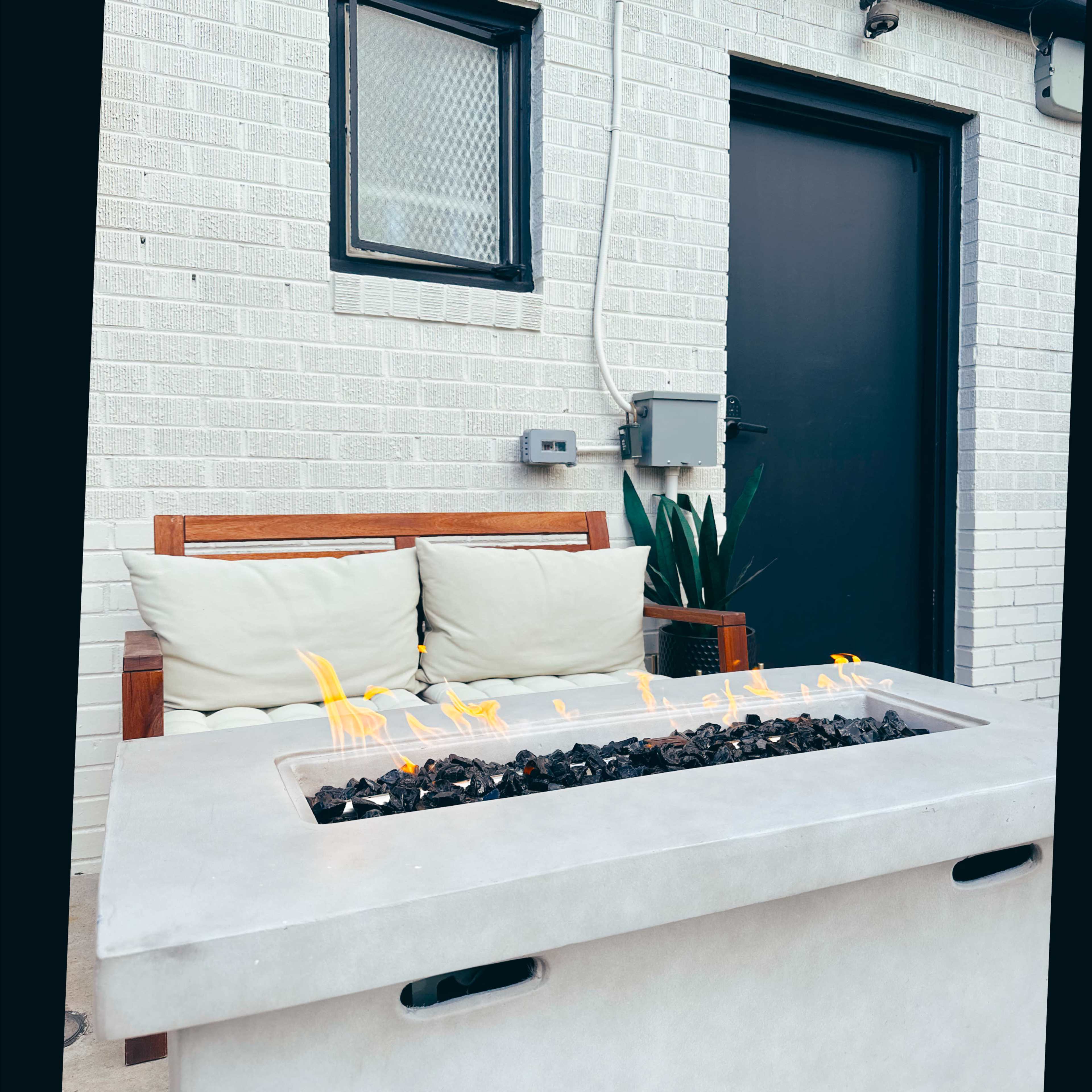 A concrete fire pit with black stones is situated next to a wooden bench with cushions against a brick wall.