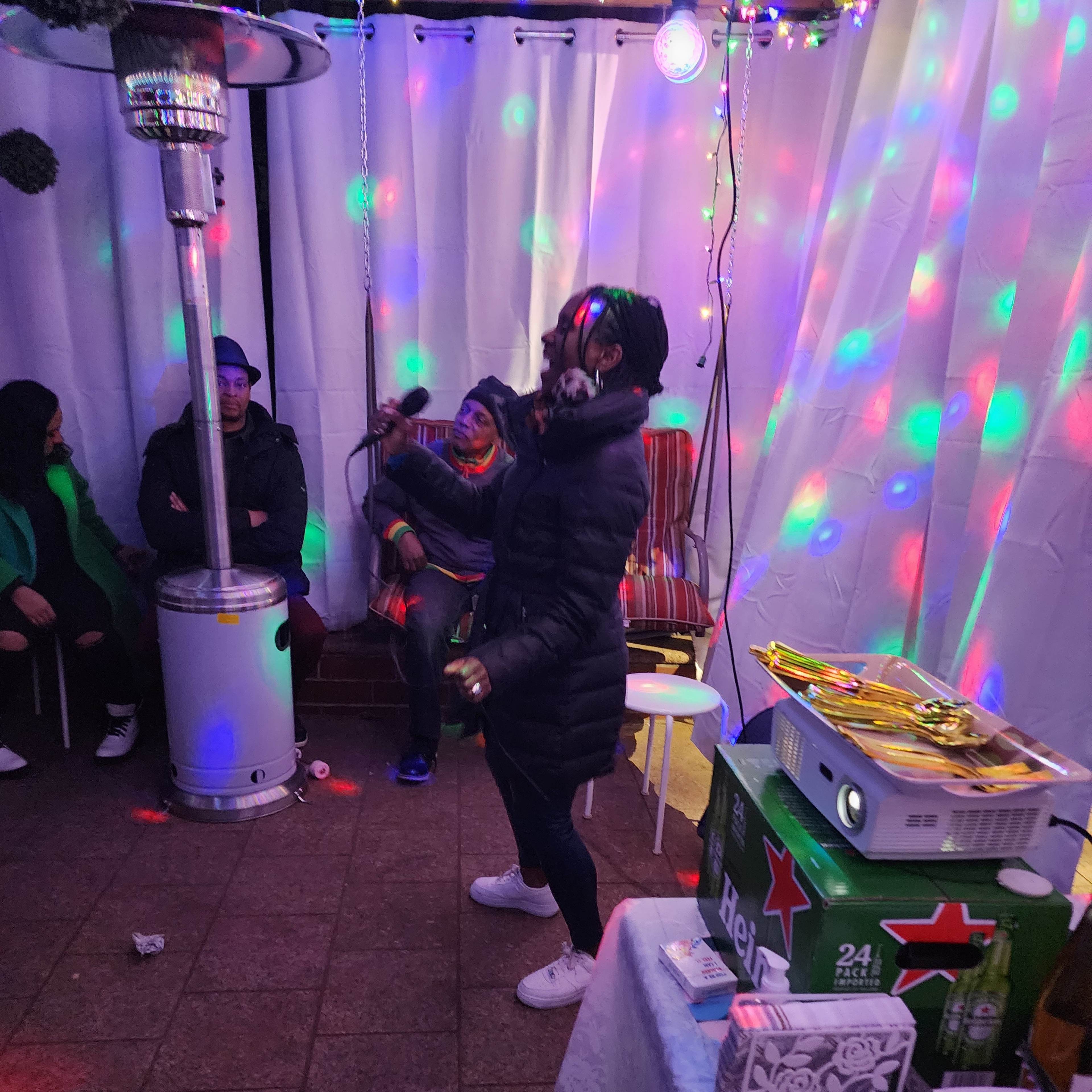 A person in a black coat dances under colorful lights in a decorated space with tables of snacks and drinks.