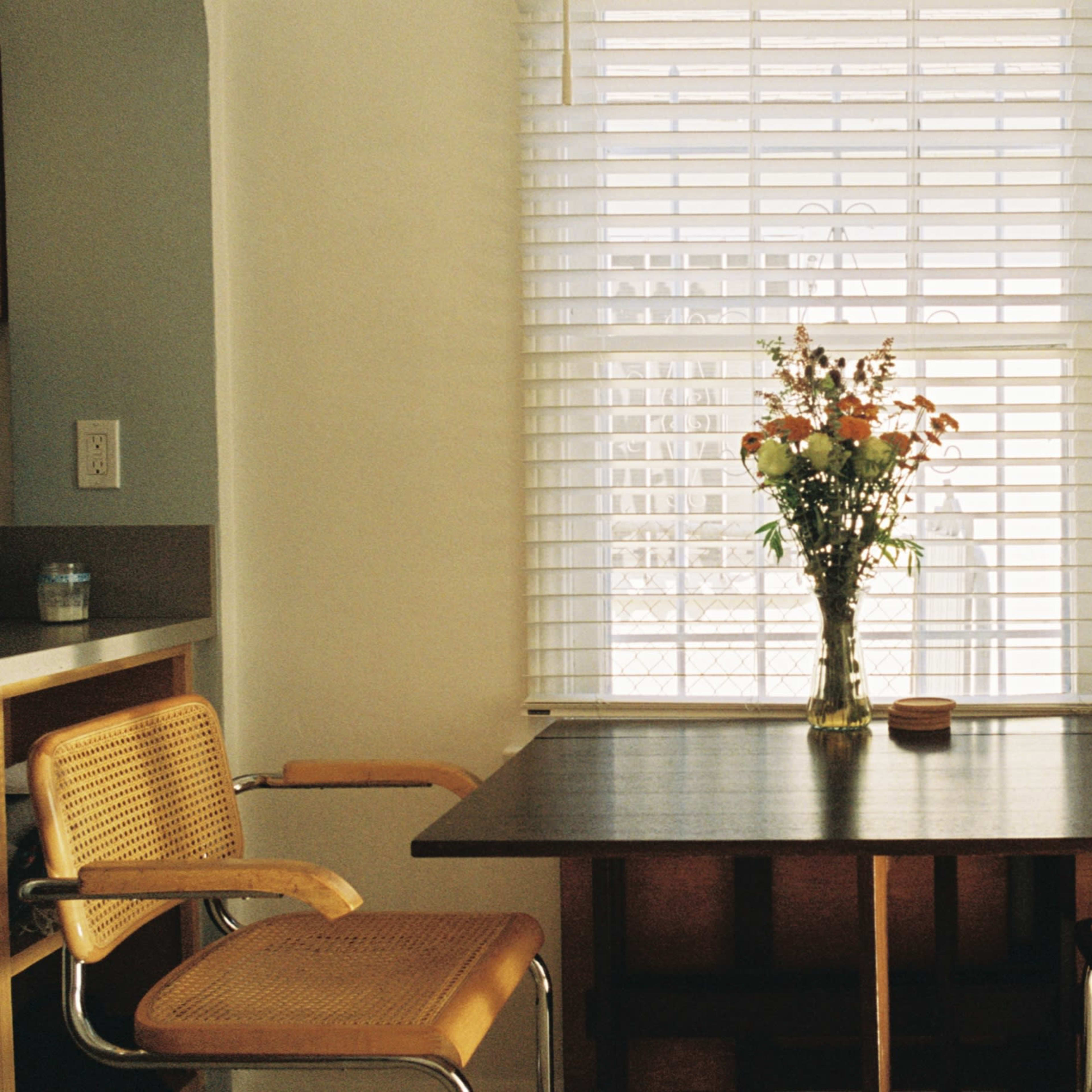 A wooden table with a vase of flowers sits between two chairs in a brightly lit room with a window covered by blinds.
