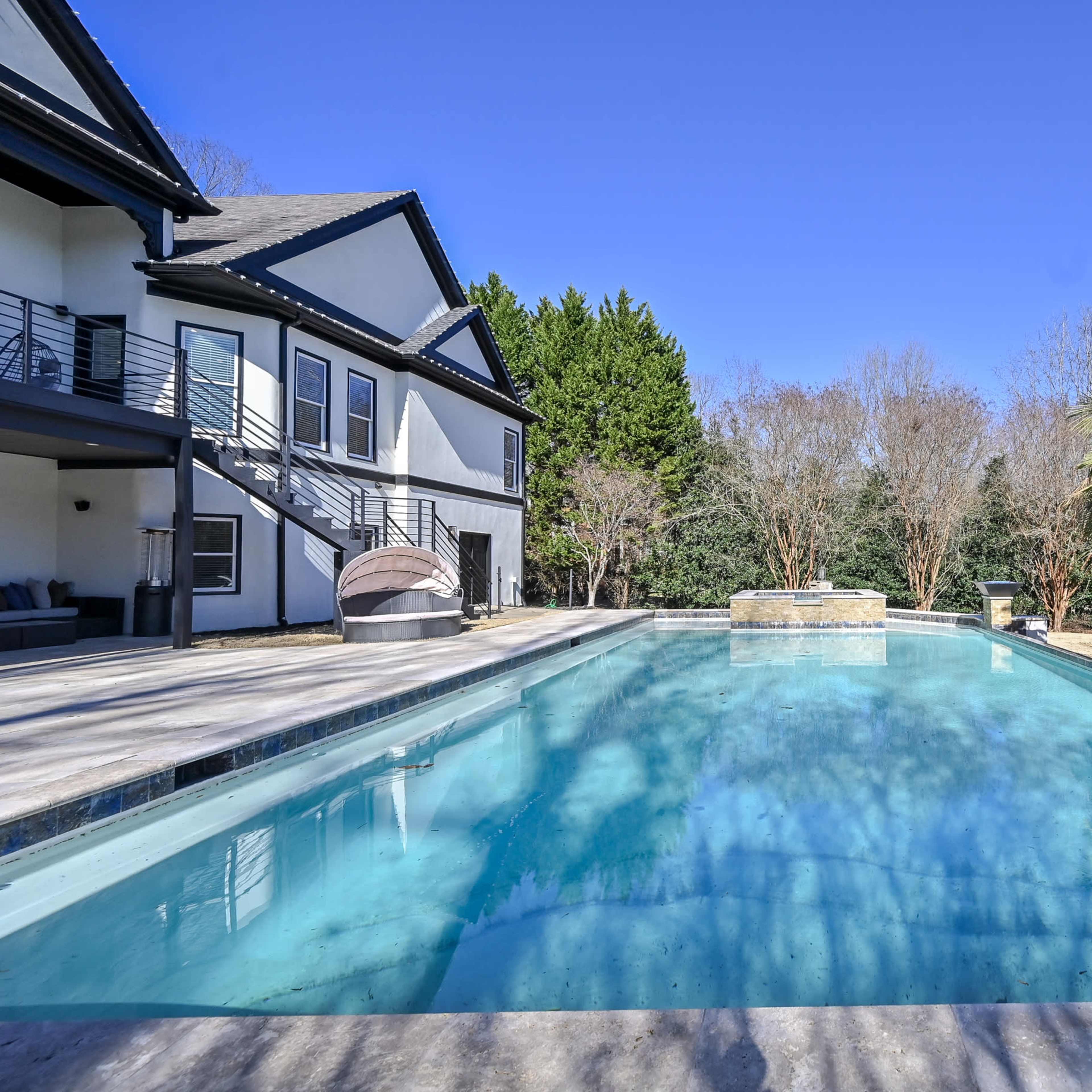 A spacious swimming pool surrounded by a large patio, with a modern house and palm trees in the background.
