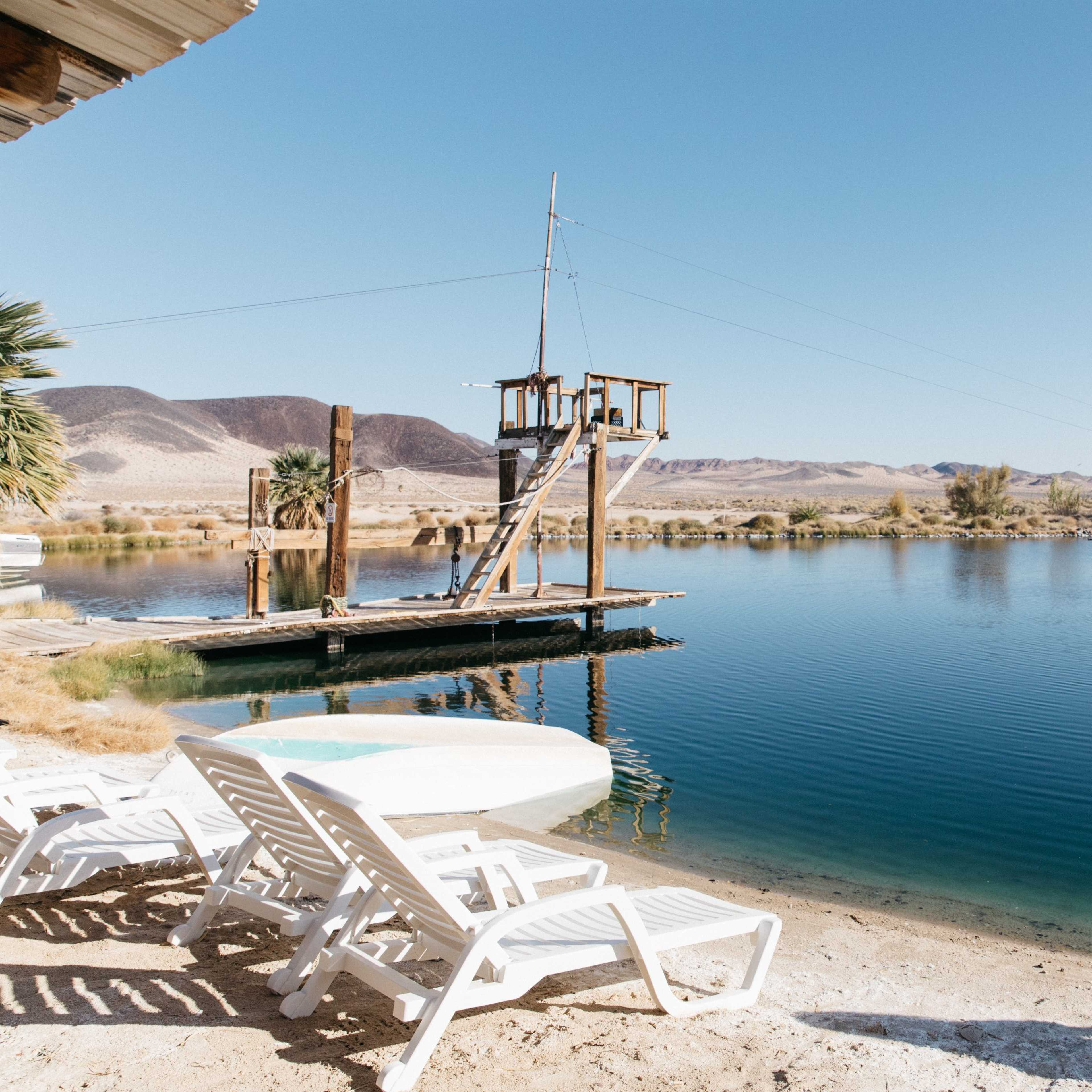 A wooden dock with a small structure extends over a calm lake surrounded by desert hills and palm trees.