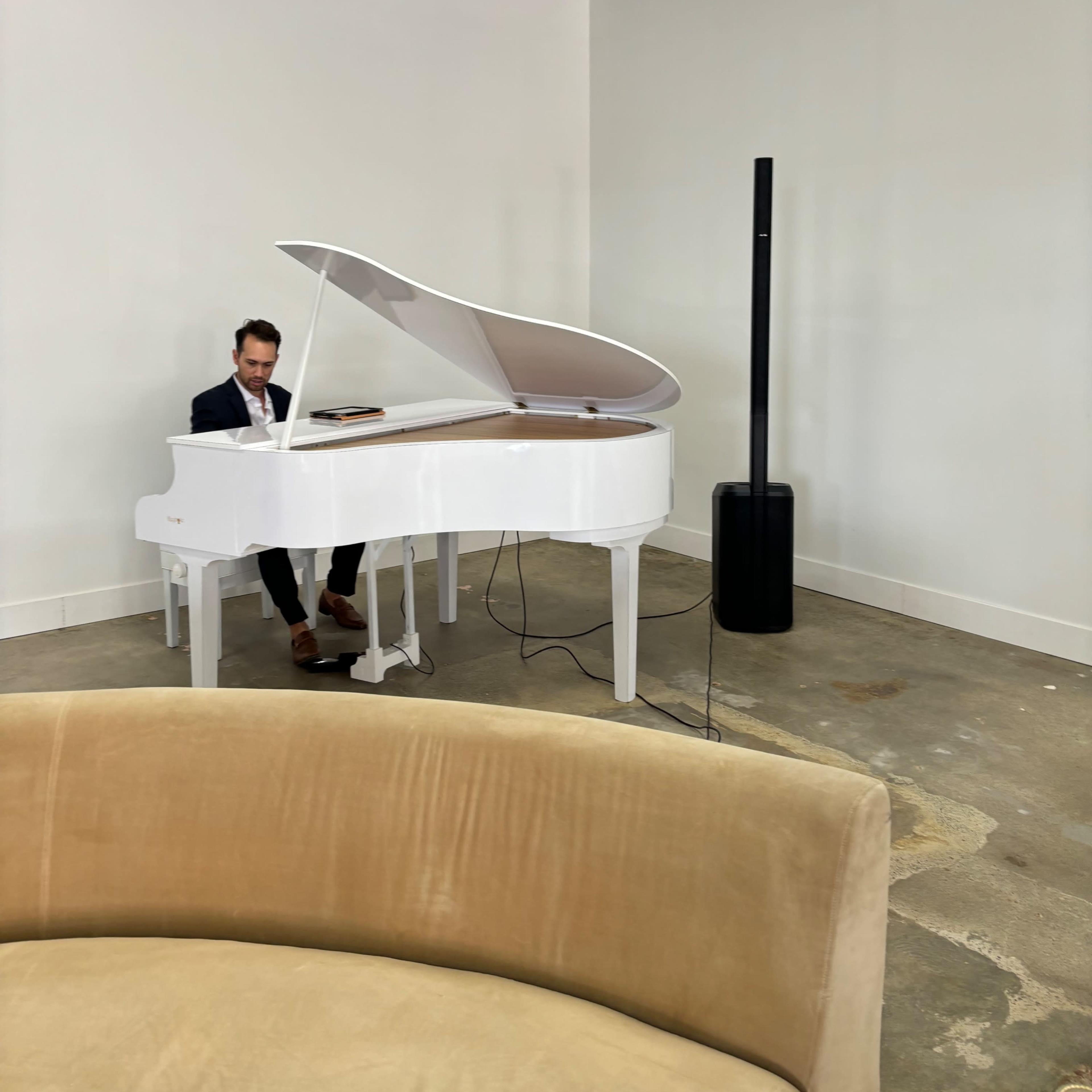 A man plays a white grand piano in a minimalist room with a beige couch in the foreground.