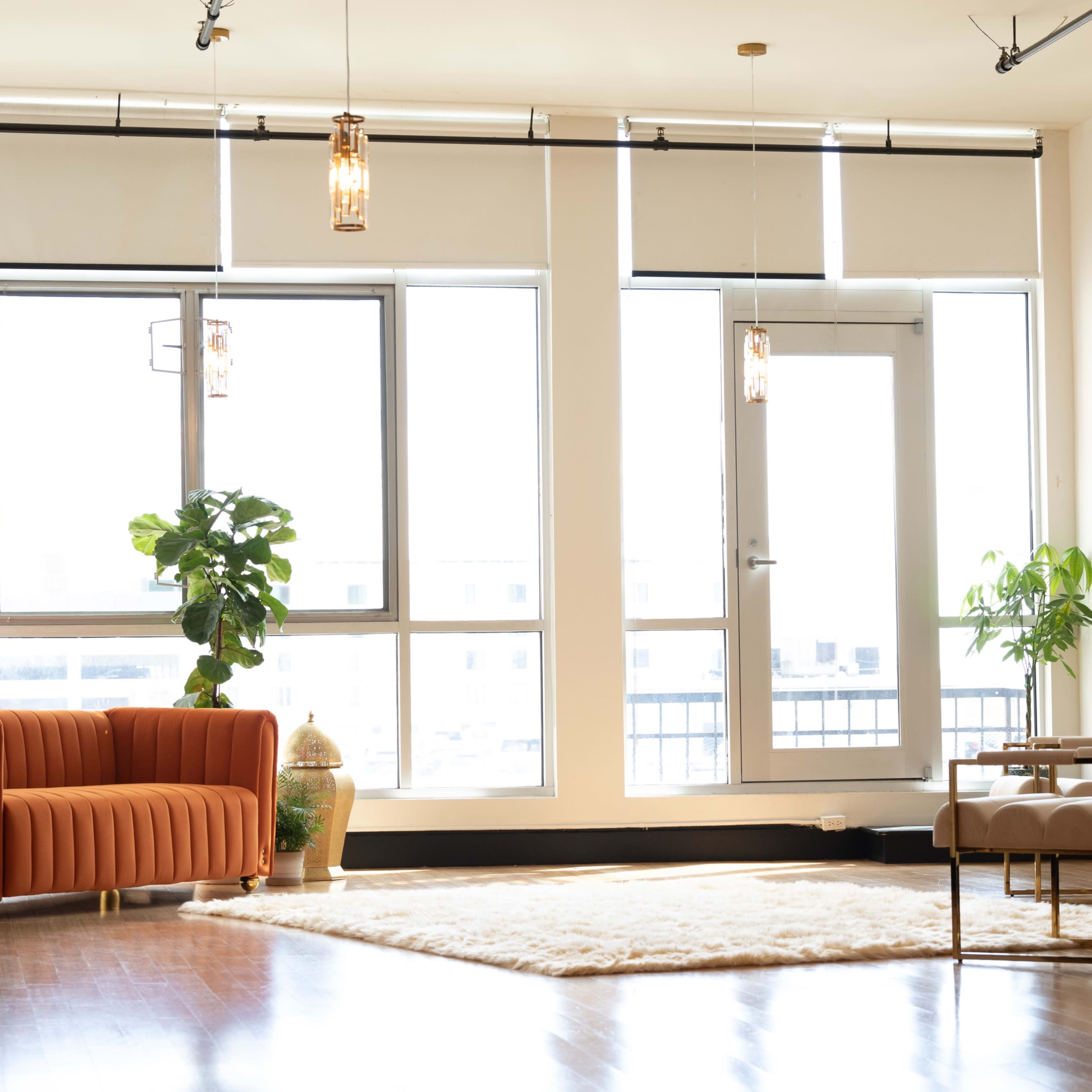 A modern living space features a rust-colored couch, a wooden chair, and several large plants near a wall of windows.