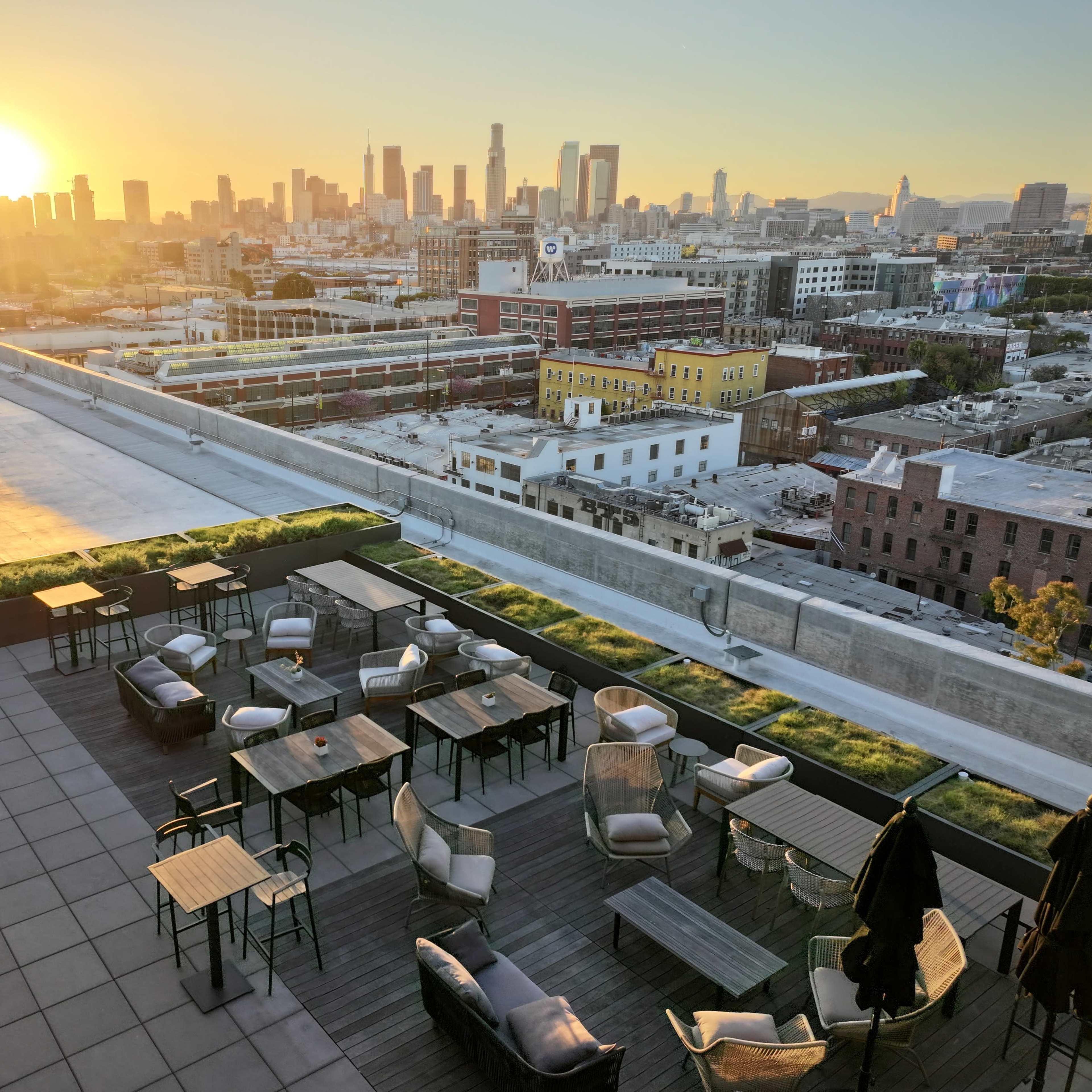 A rooftop terrace with wooden tables and chairs overlooking a city skyline at sunset.
