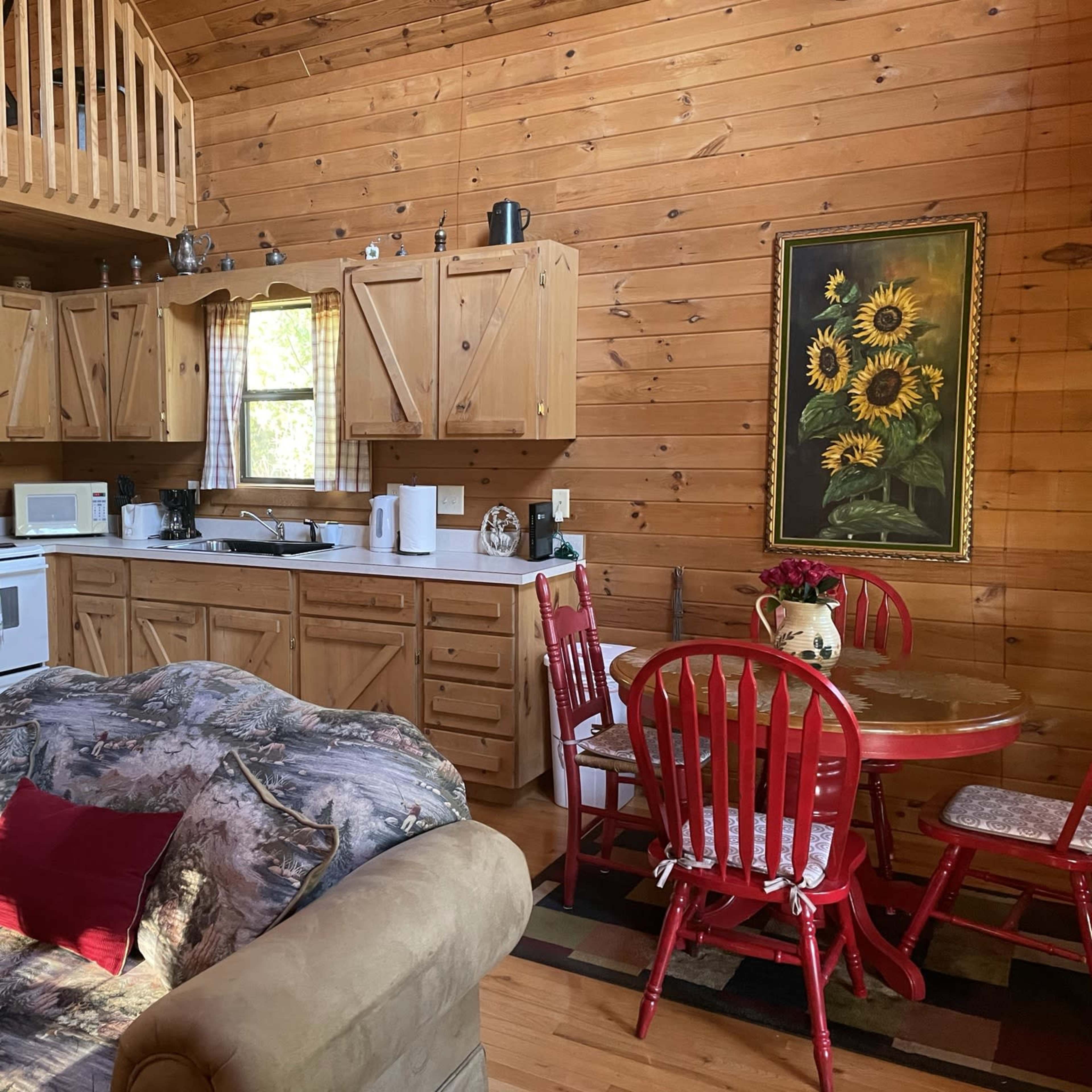 A wooden cabin interior features a kitchen area with pine cabinets, a white refrigerator, and a dining table with red chairs.