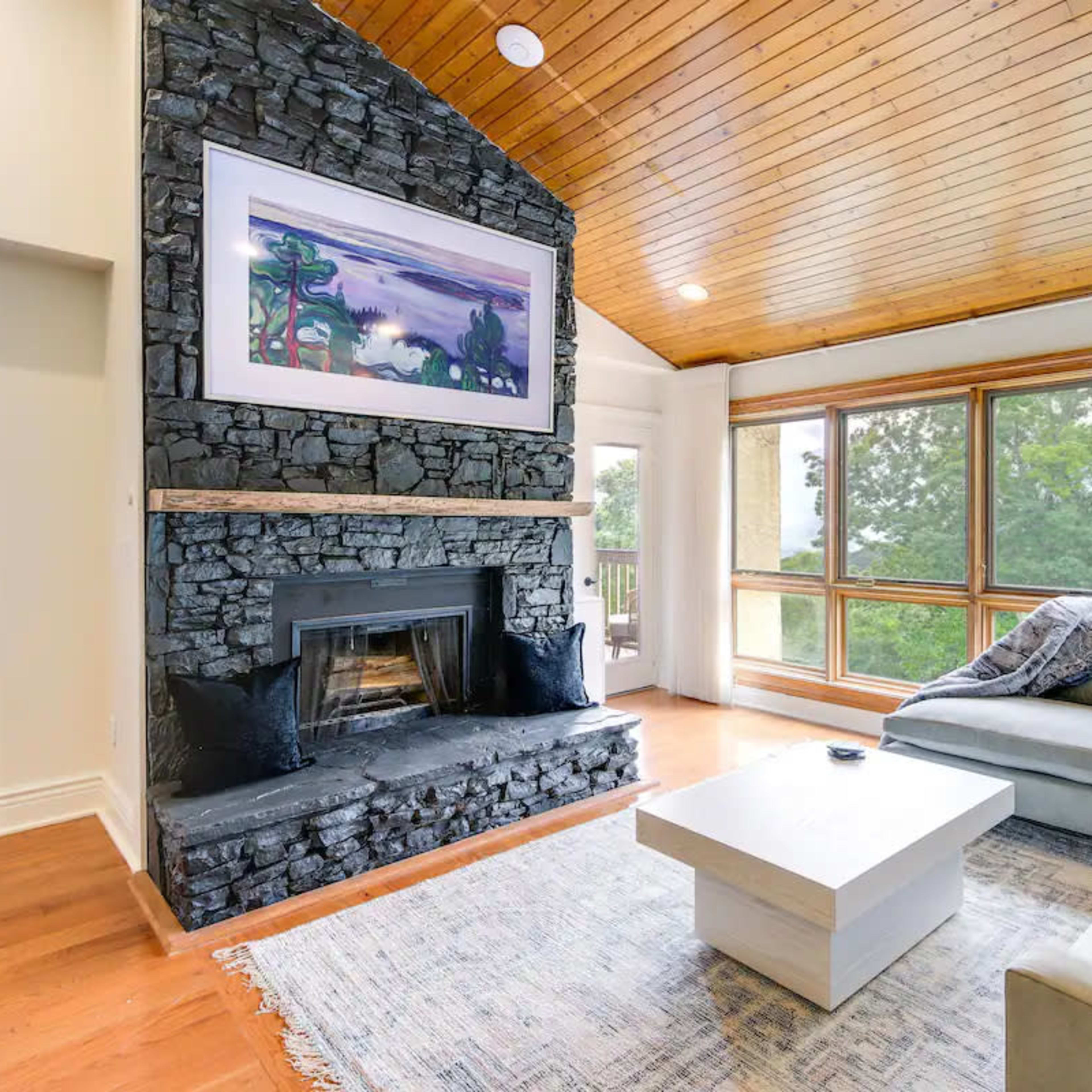 A living room with a stone fireplace, a sloped wooden ceiling, large windows, and a light-colored sofa facing a coffee table.