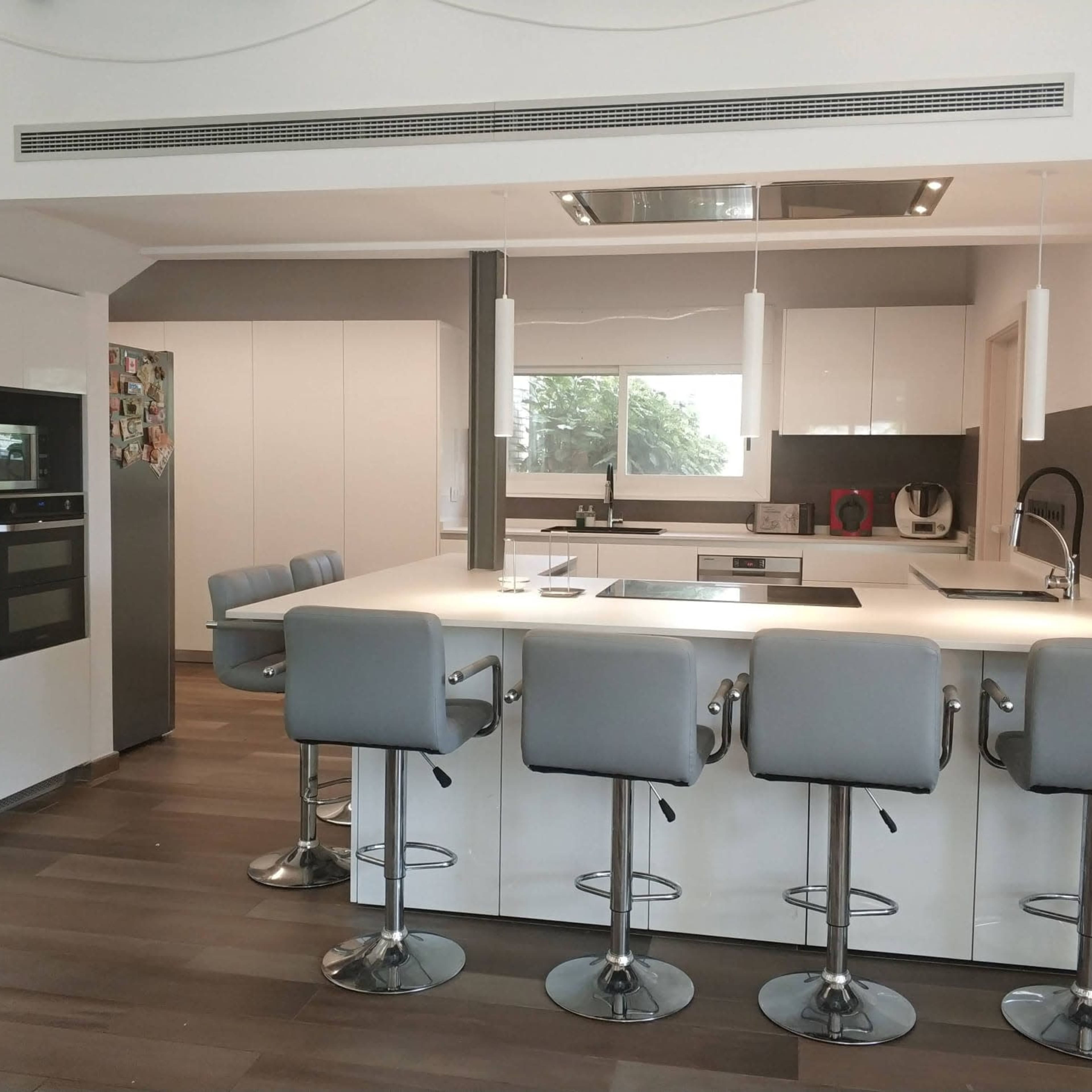 A modern kitchen features a central island with four gray bar stools and sleek cabinetry, with a view through a window to the outside.