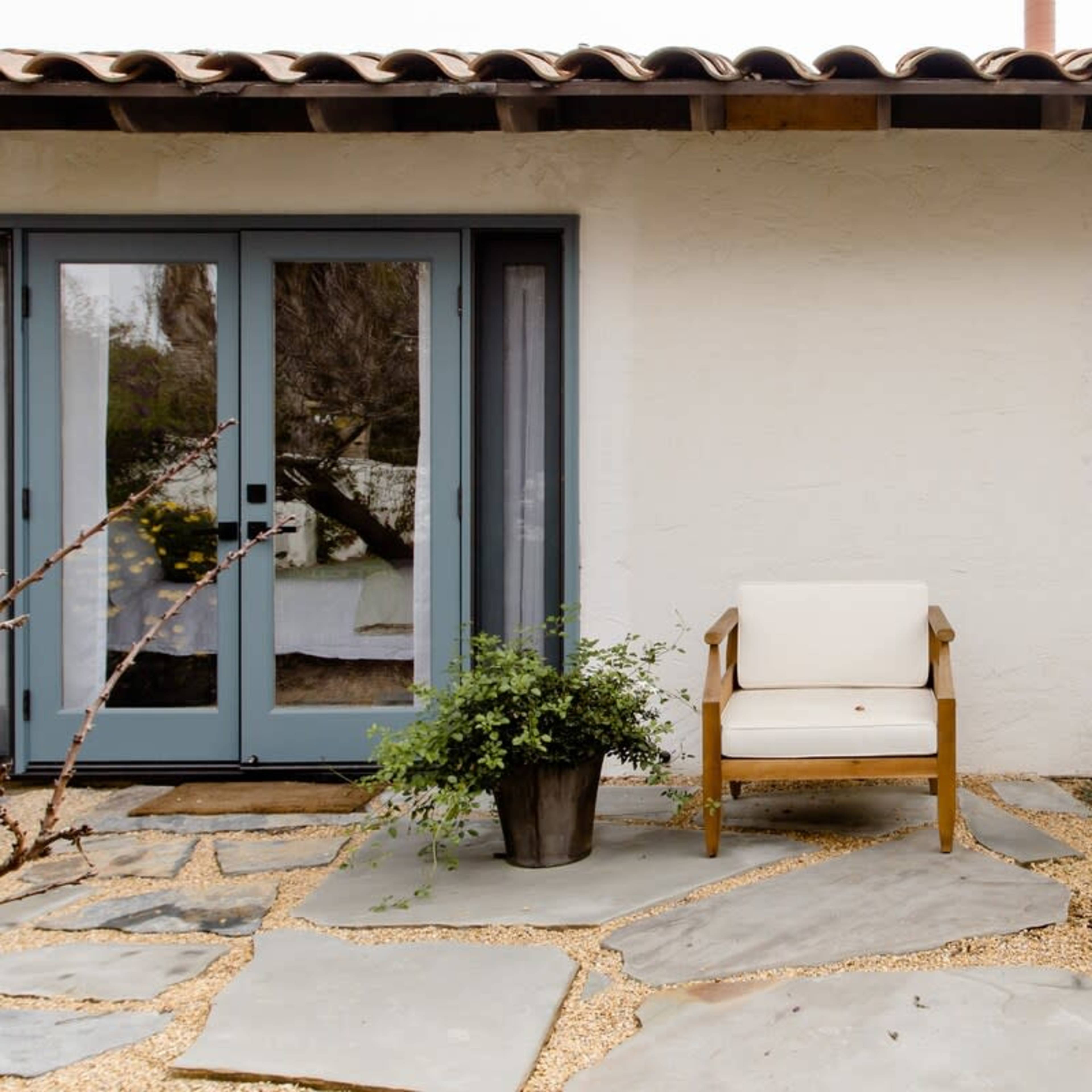 A wooden chair with a white cushion is placed on a stone pathway next to a house with light-colored walls and blue doors.