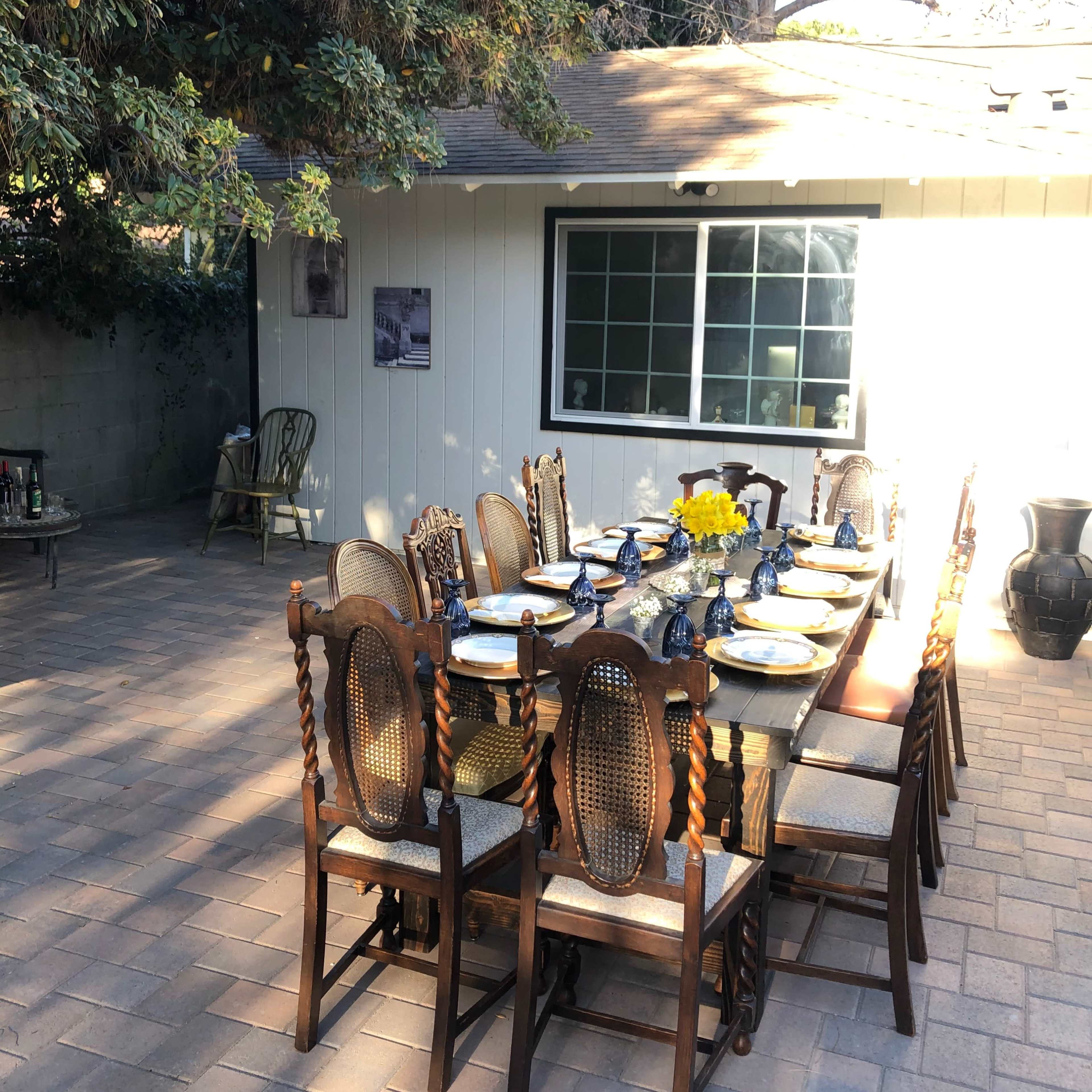 A long dining table is set with dishes and flowers in an outdoor space next to a house.