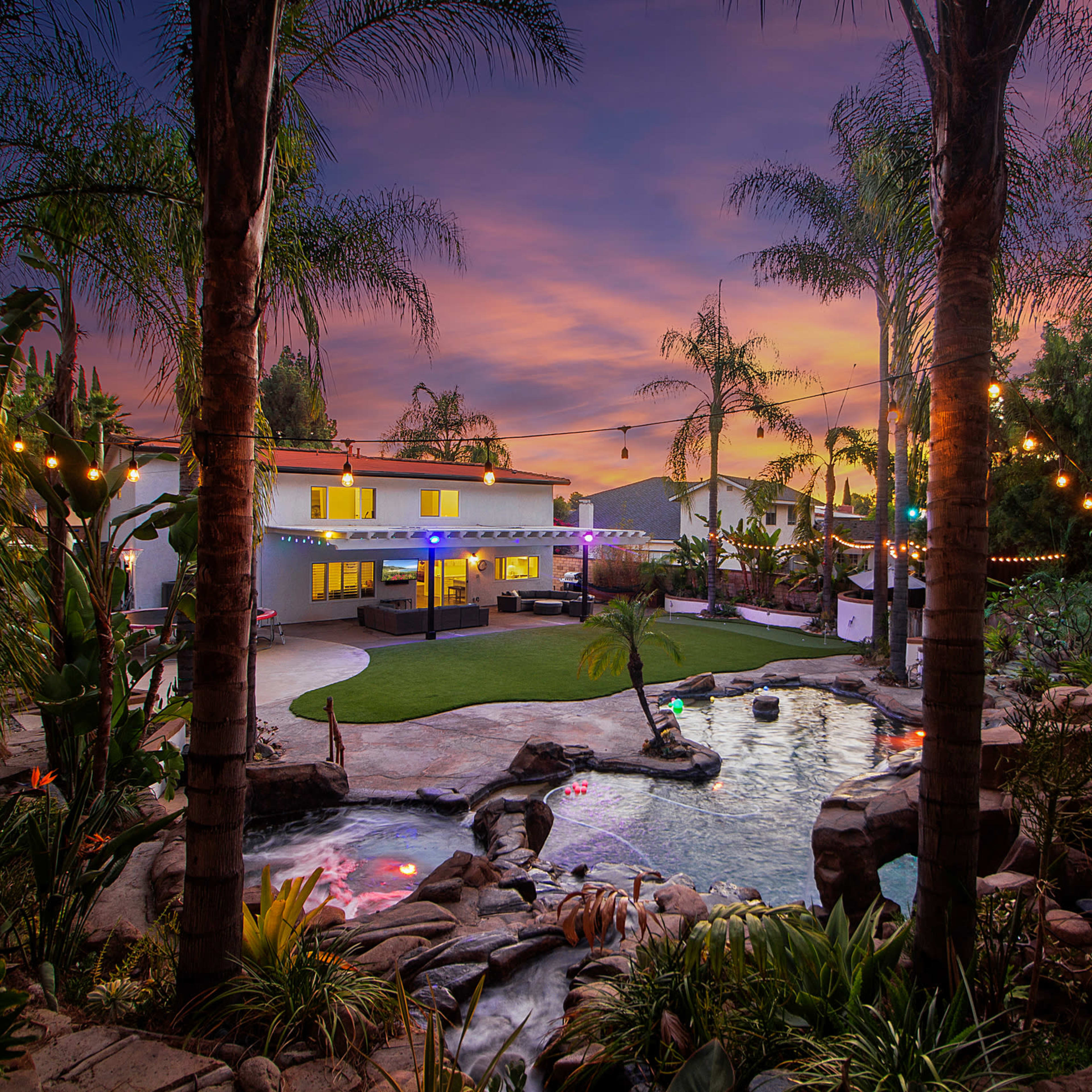 A backyard scene features a modern home surrounded by palm trees, colorful lights, and a landscaped area with a pool and waterfall at sunset.