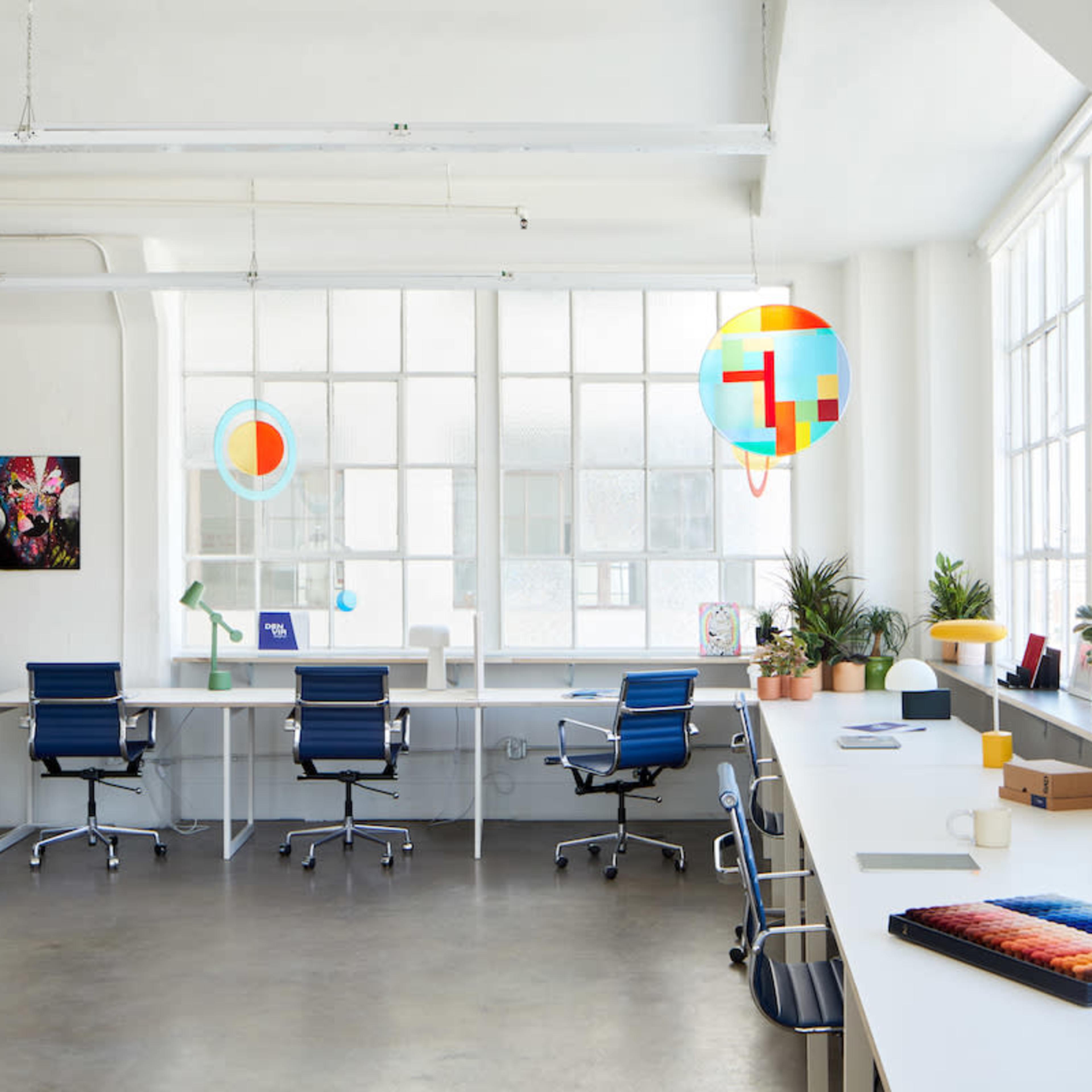 A bright, modern office space with large windows, a few desks with chairs, and various decorative plants.