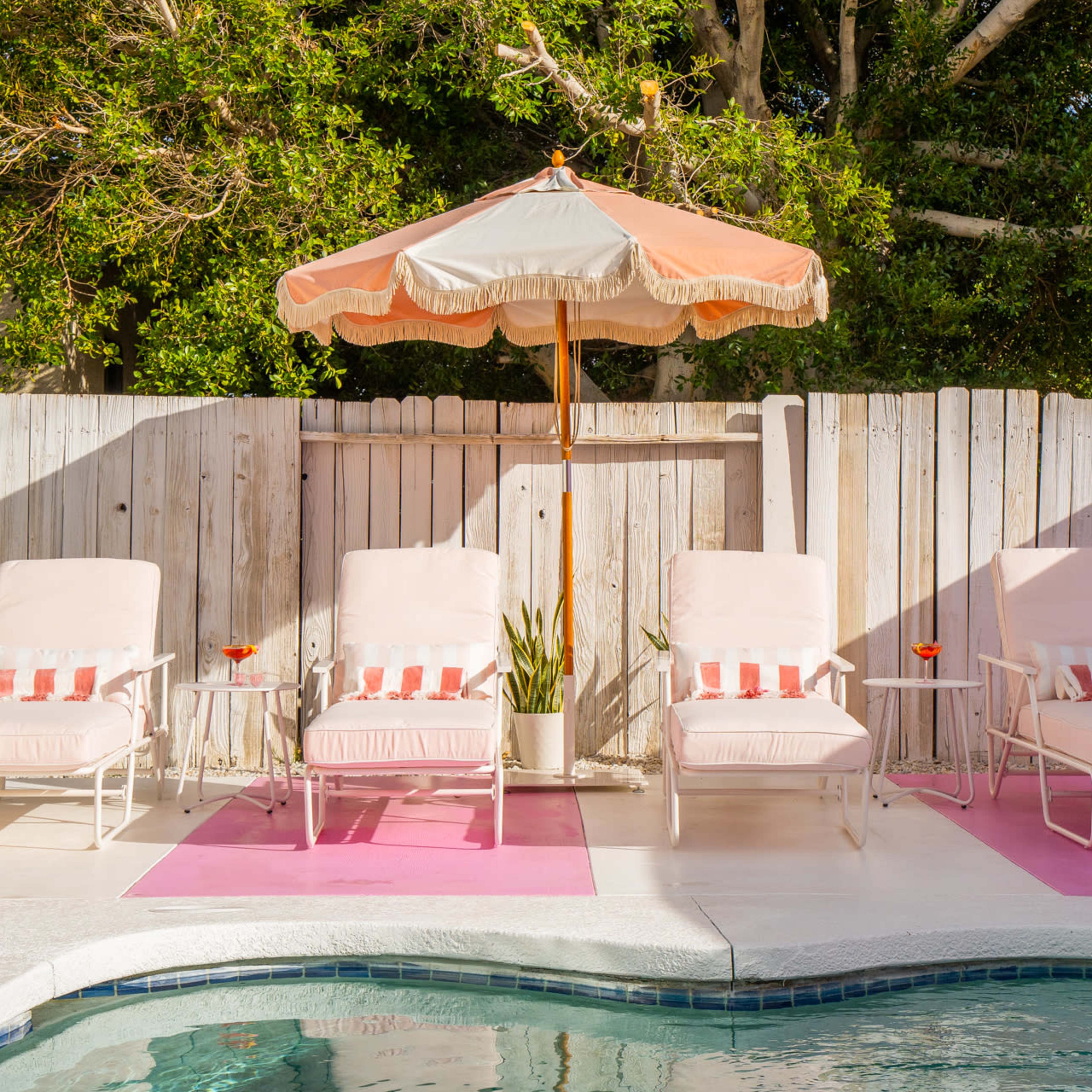 A row of four lounge chairs with white cushions and pink striped pillows is arranged beside a swimming pool, shaded by a large orange and cream umbrella.