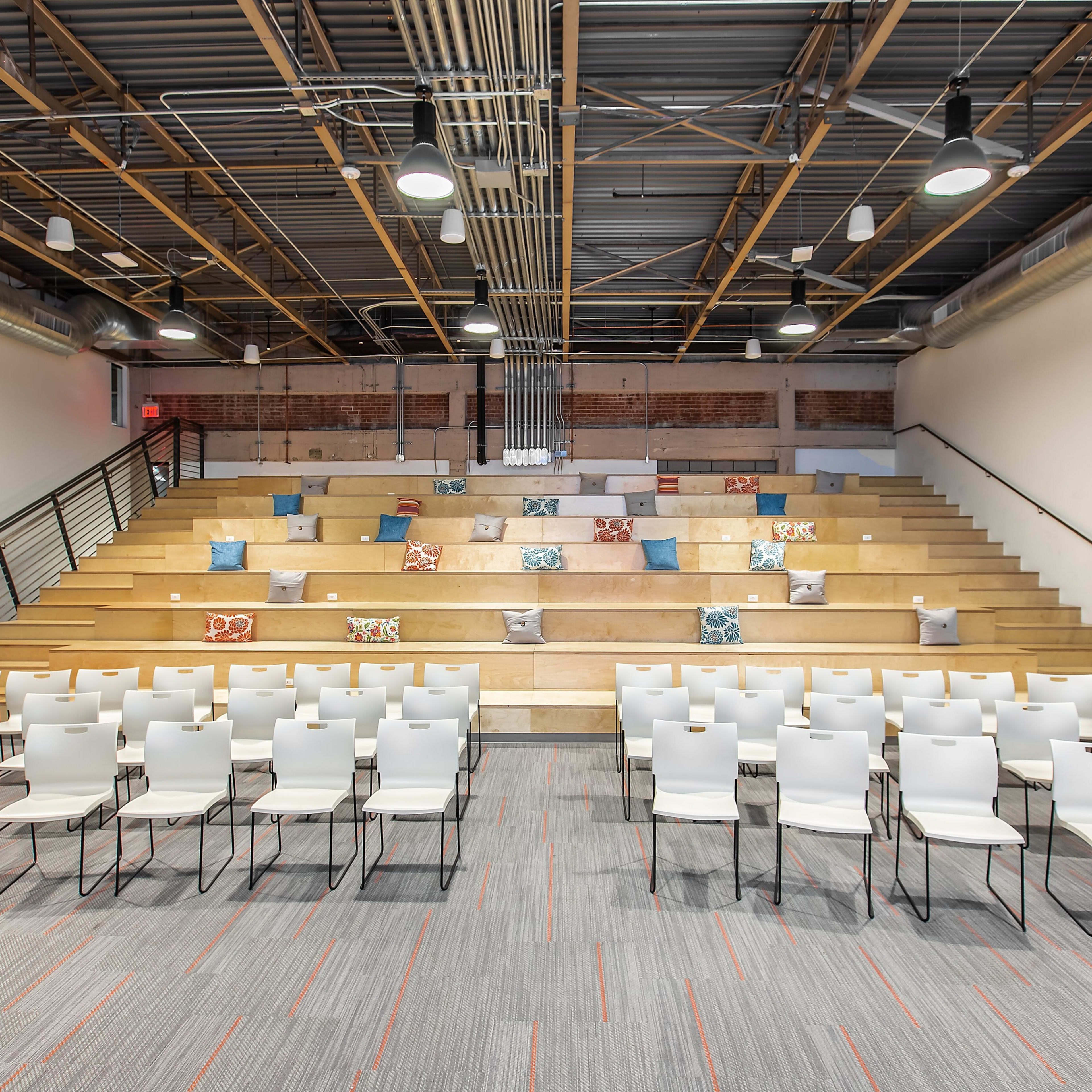A modern auditorium setting with tiered seating, white chairs arranged neatly in rows, and decorative pillows placed on the steps.