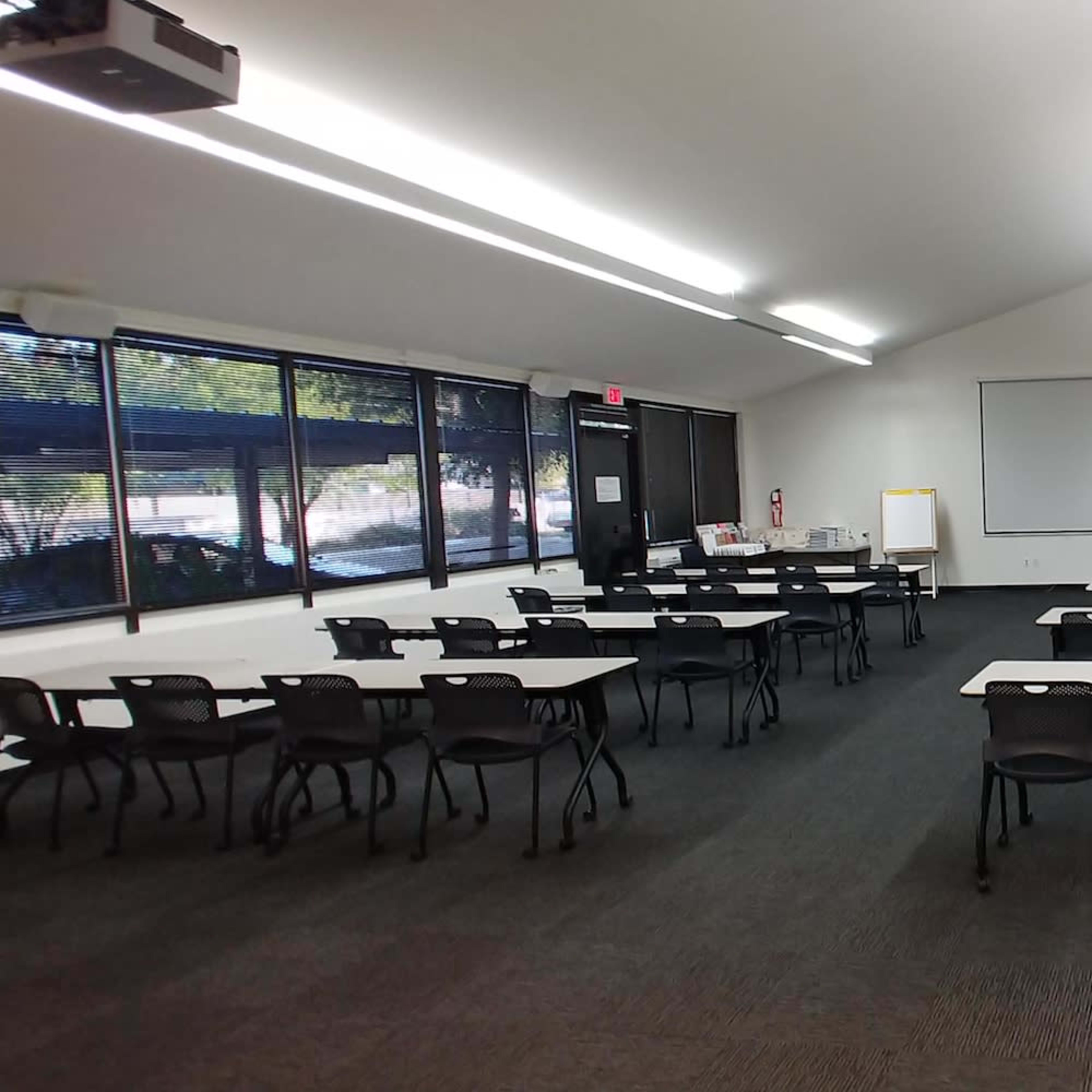 A spacious classroom is arranged with rows of tables and chairs, featuring large windows that allow natural light to enter.