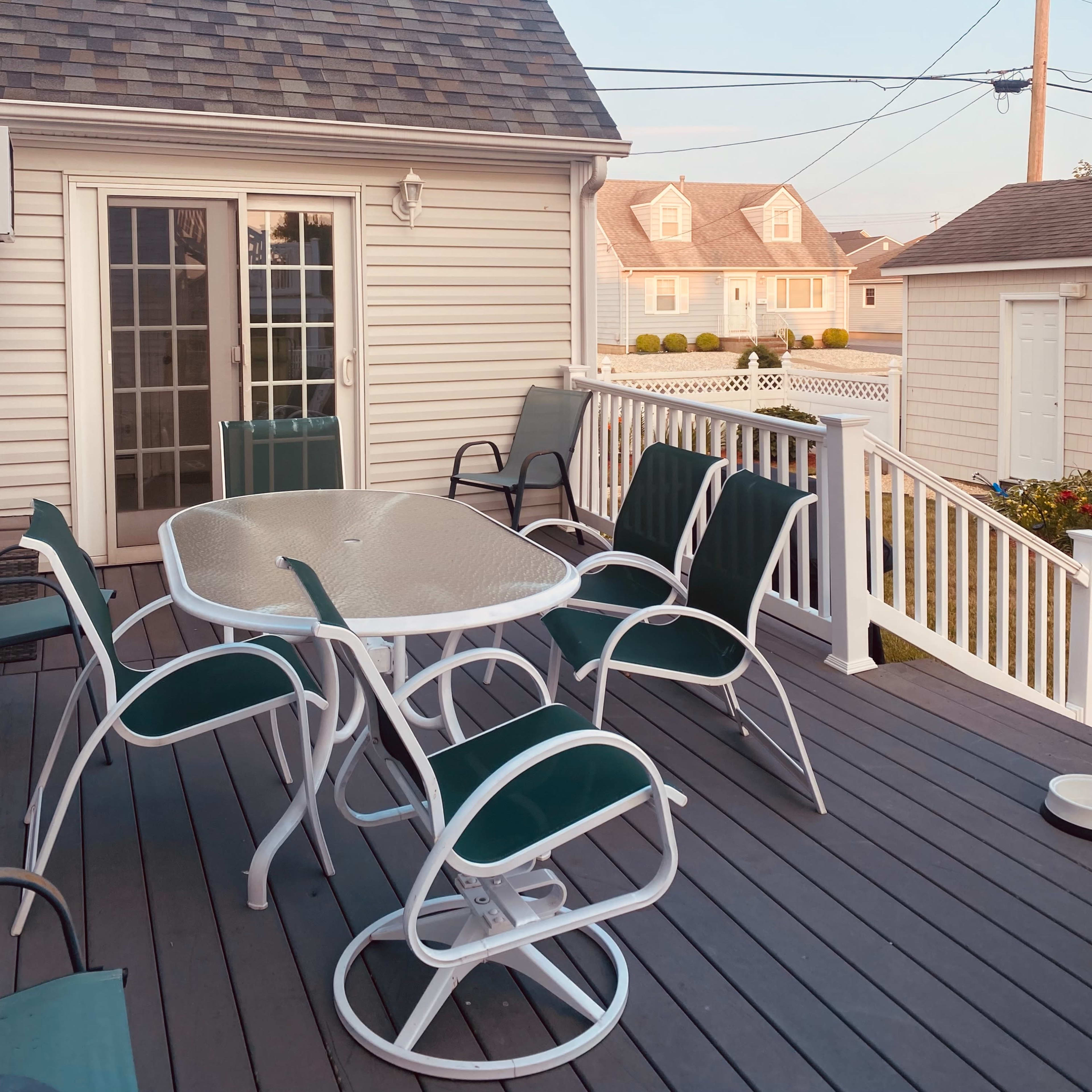 A deck with a round table and green chairs overlooks nearby houses and power lines under a clear sky.