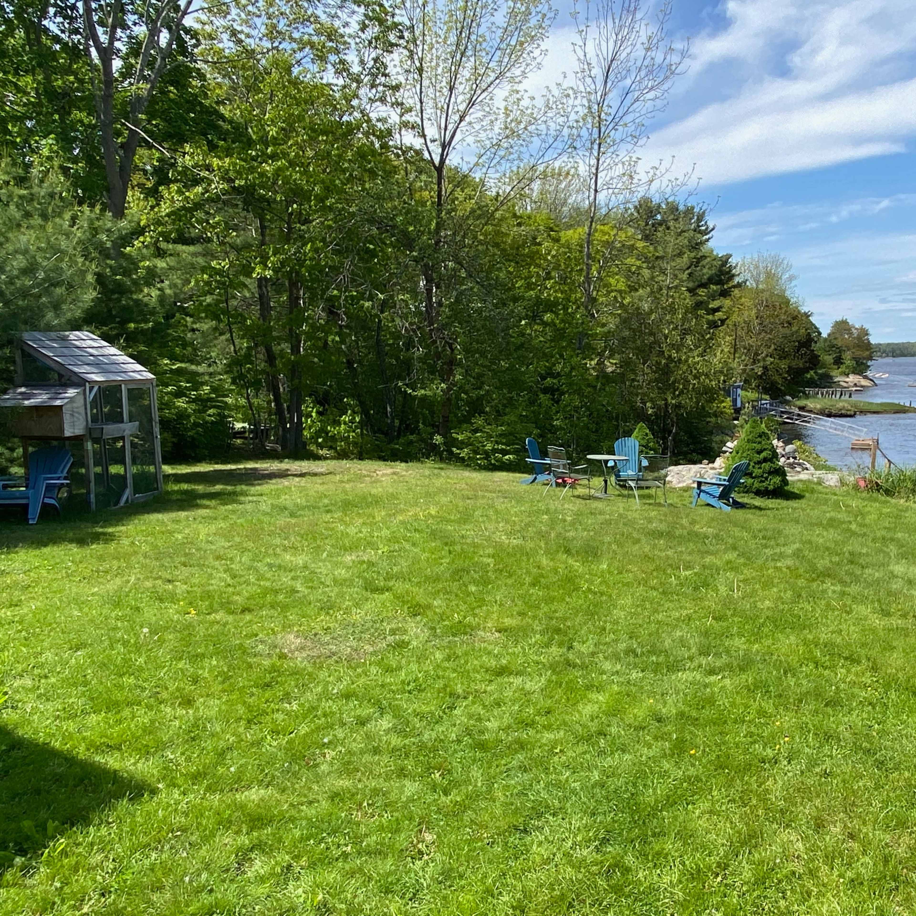 A grassy area near a river features blue Adirondack chairs and a small shed, with trees lining the shore under a clear sky.