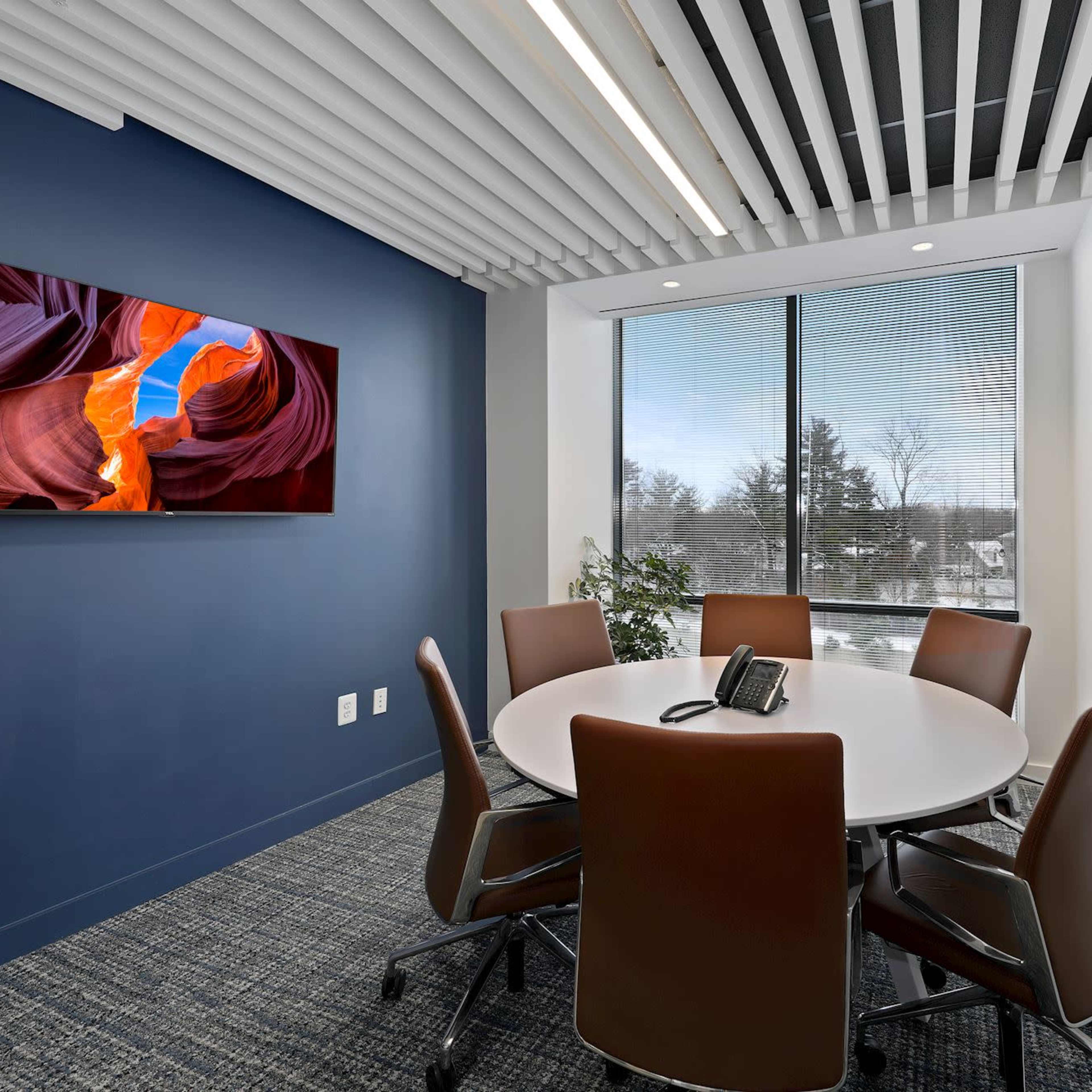 A modern conference room features a round table with six chairs, a wall-mounted TV, and a large window overlooking a exterior landscape.
