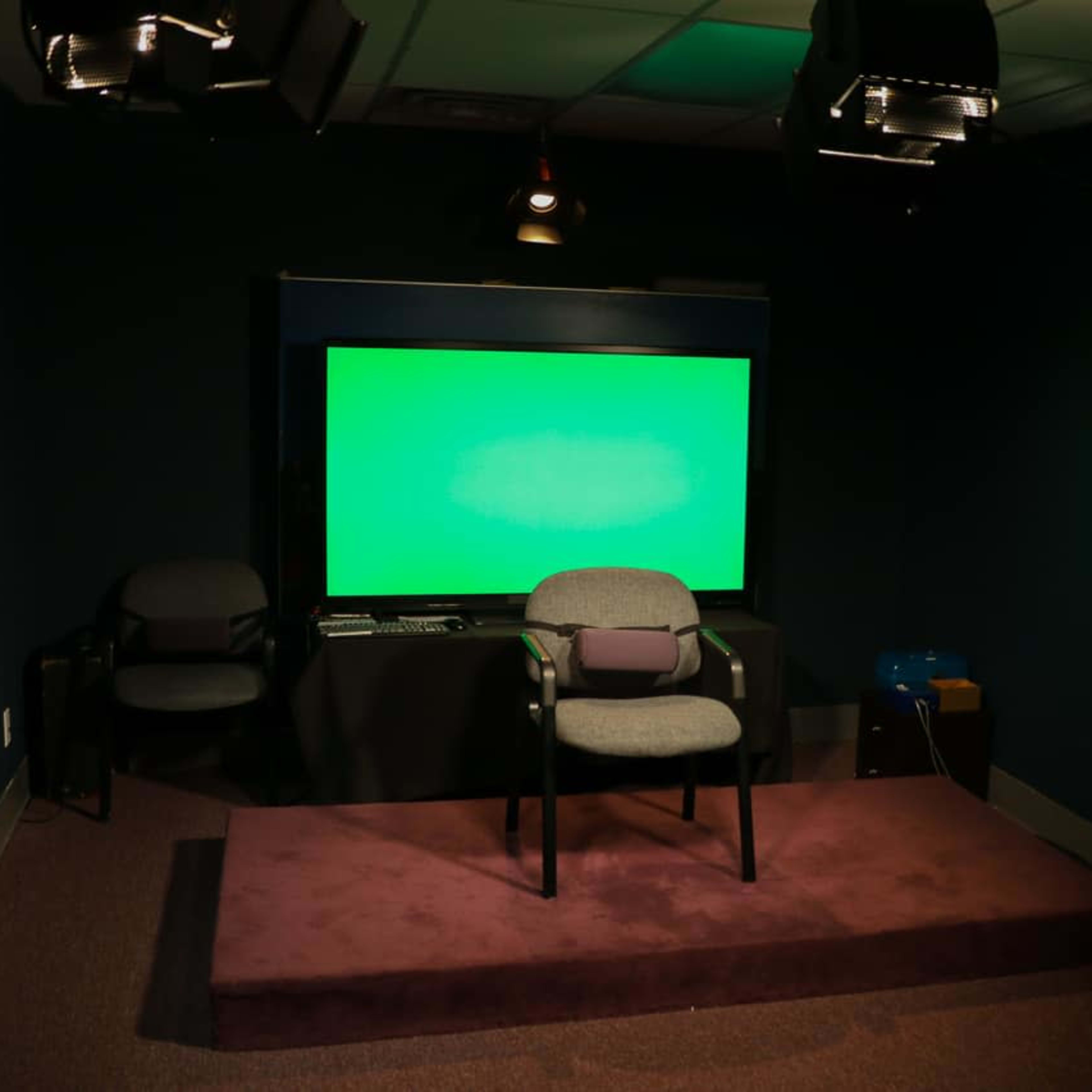 A dimly lit studio with a green screen, a chair on a raised platform, and various lighting equipment.