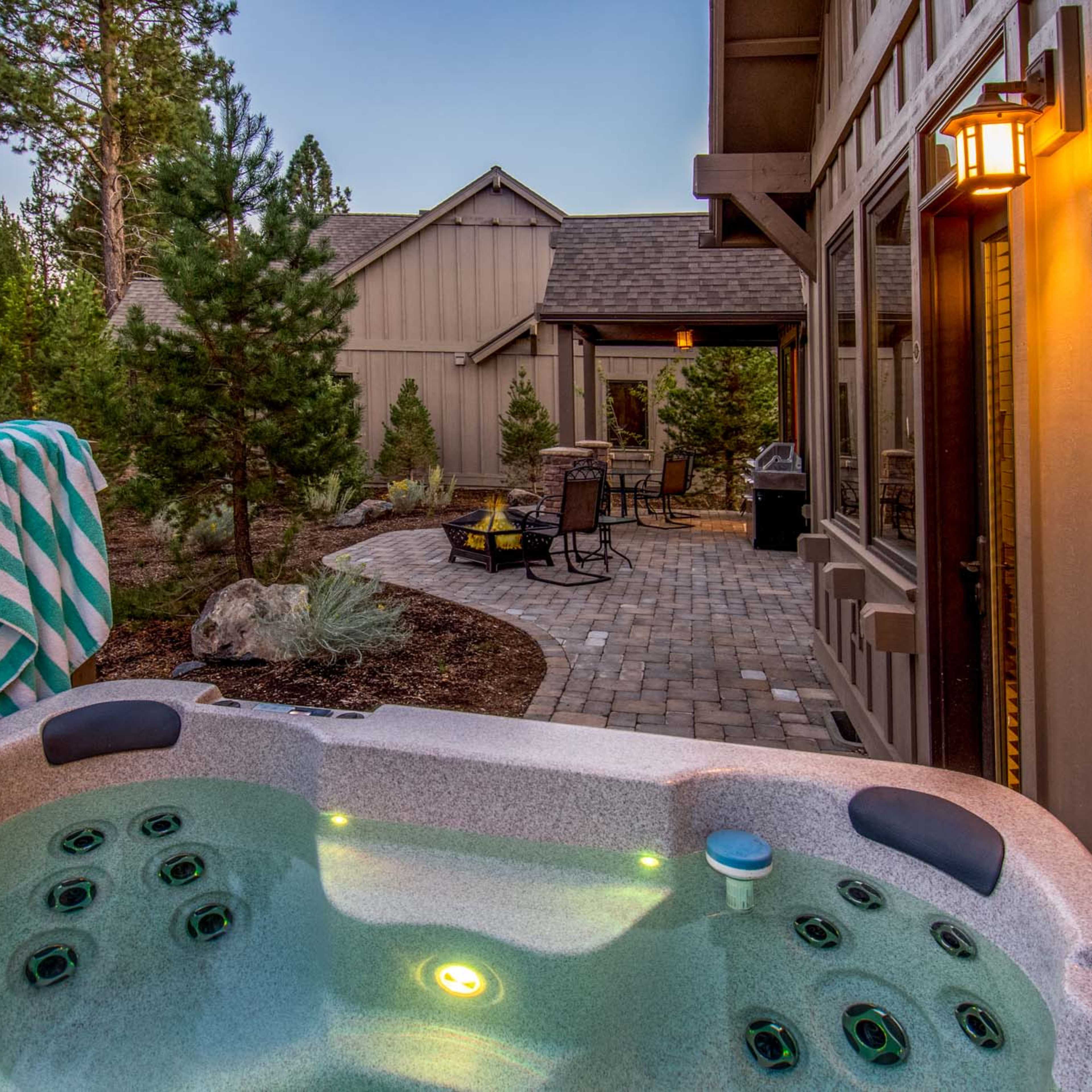 A hot tub is positioned on a patio, surrounded by trees and a stone walkway leading to a house with outdoor seating and a grill.