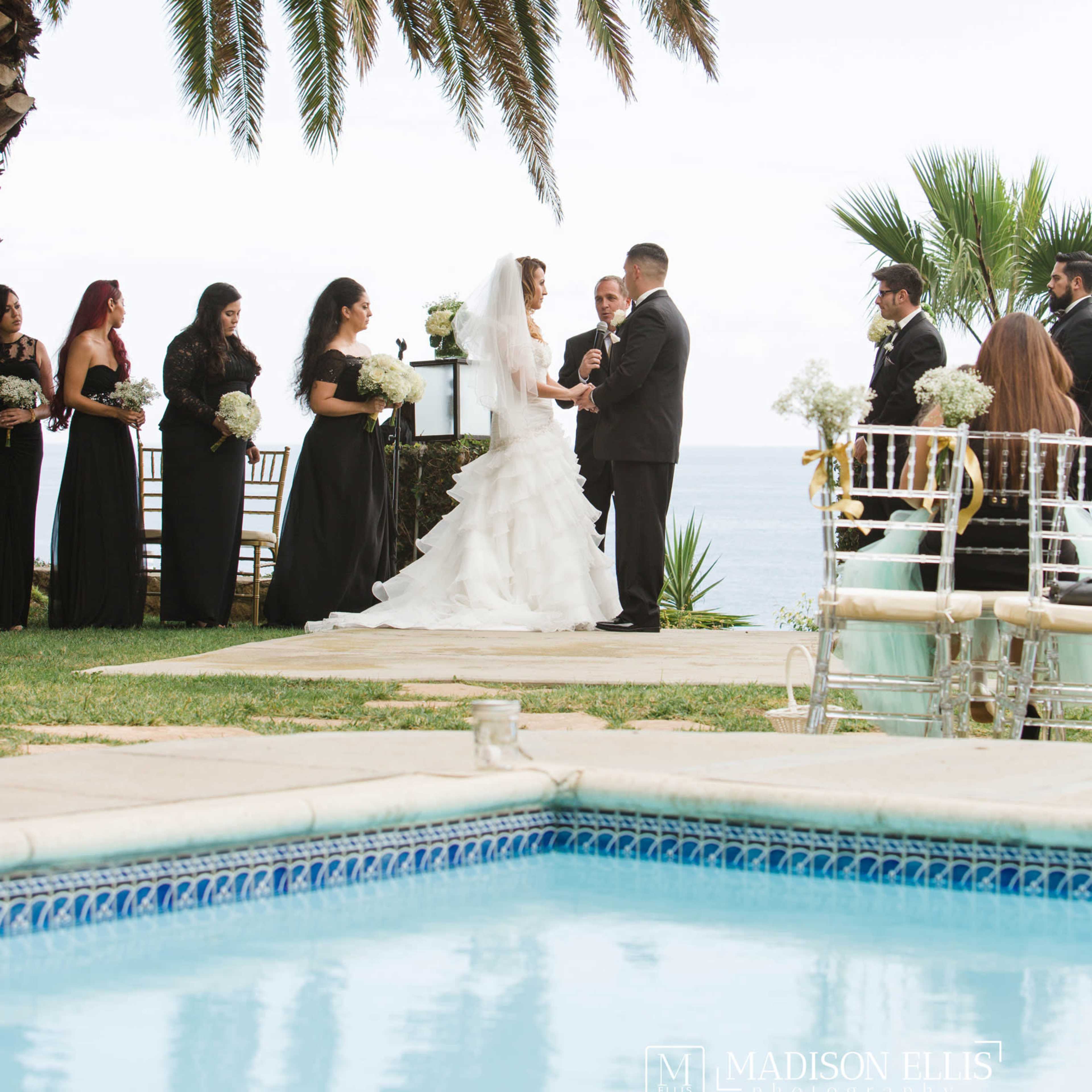 A couple exchanges vows at an outdoor wedding ceremony near a swimming pool, with guests in black attire seated around them.