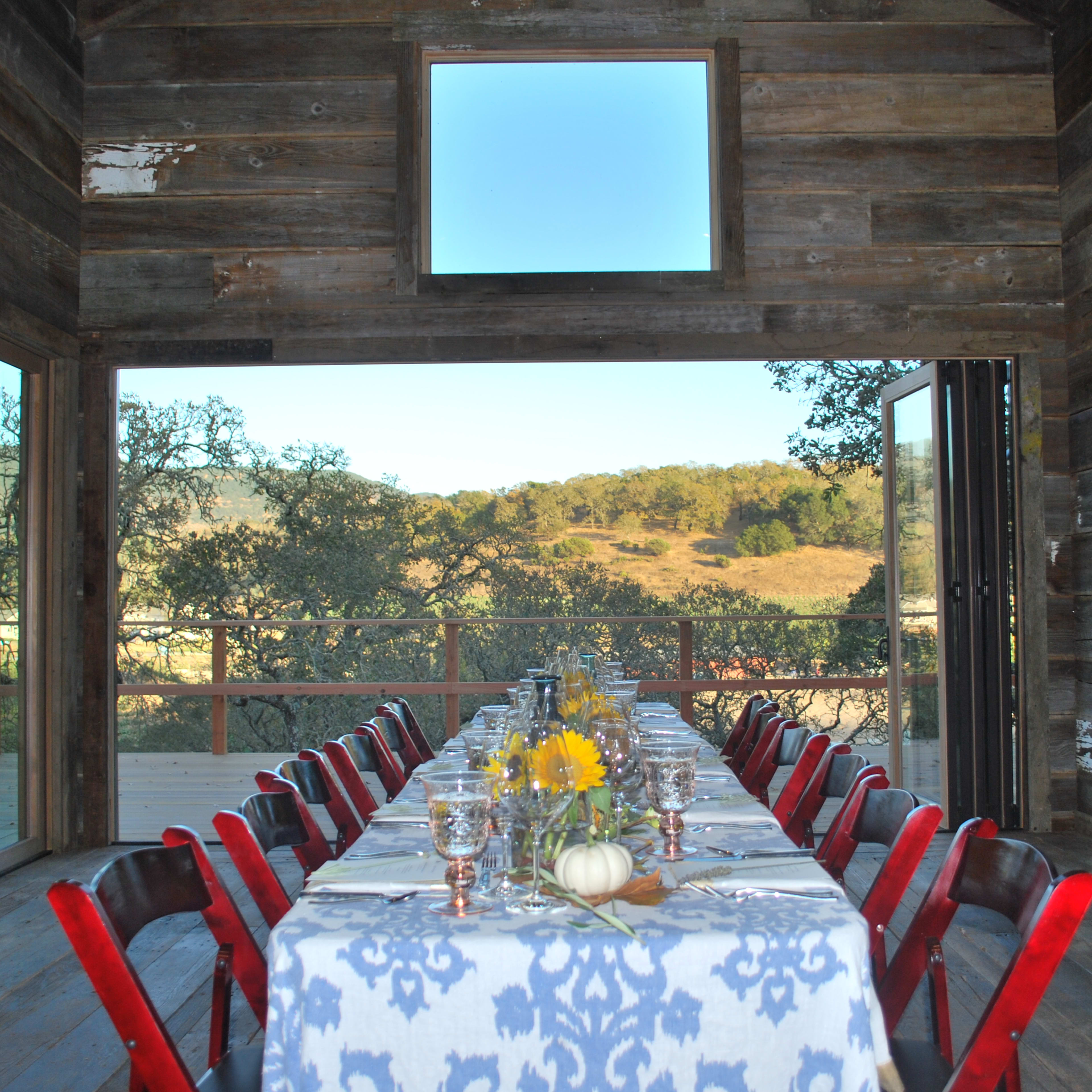 A large dining table is set with a blue and white tablecloth inside a rustic wooden room, overlooking a scenic outdoor landscape with rolling hills and trees.