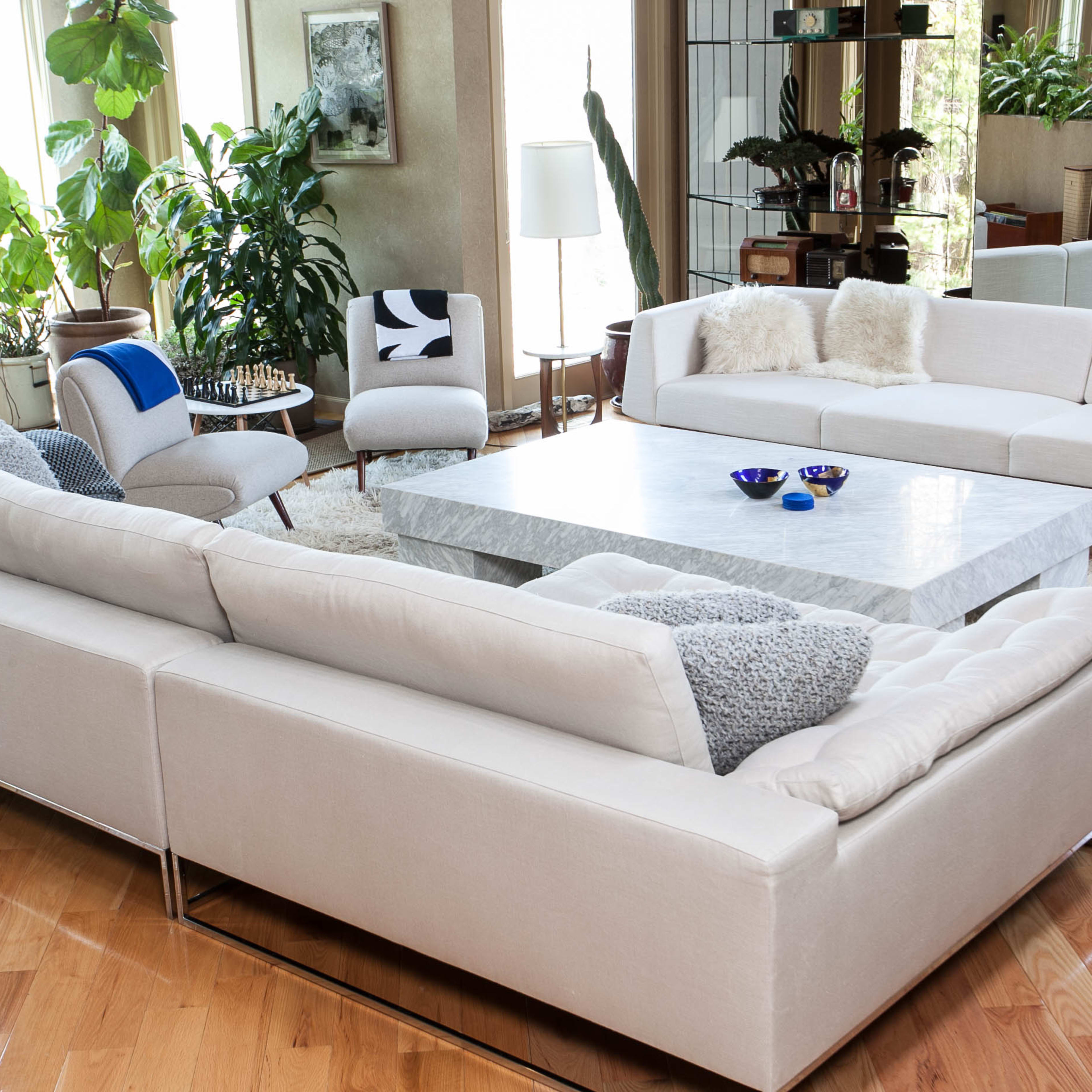 The living room features a spacious layout with a large stone coffee table surrounded by a light-colored sectional sofa and contemporary chairs, all set among various indoor plants.