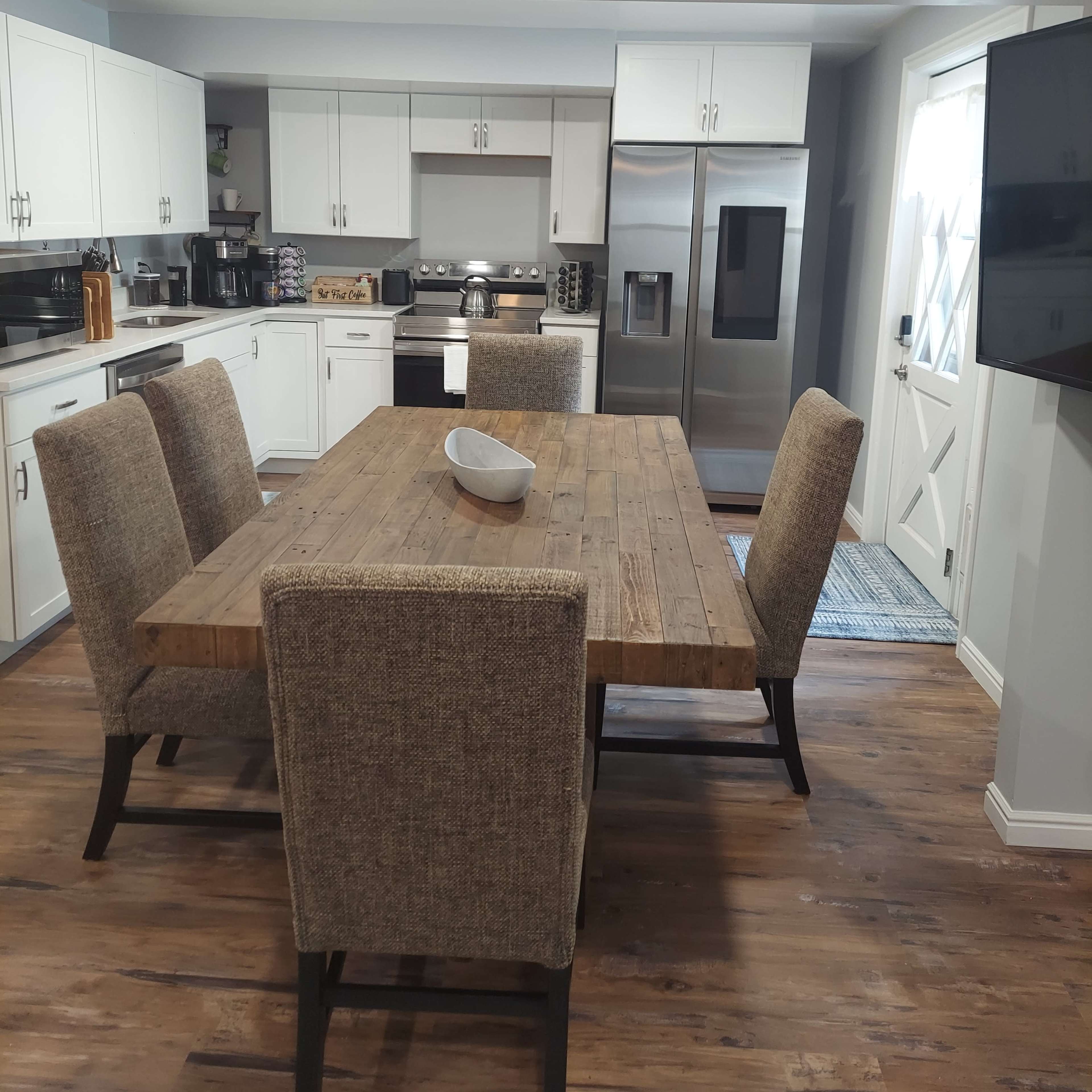 A modern kitchen with a large wooden dining table surrounded by upholstered chairs, stainless steel appliances, and white cabinetry.