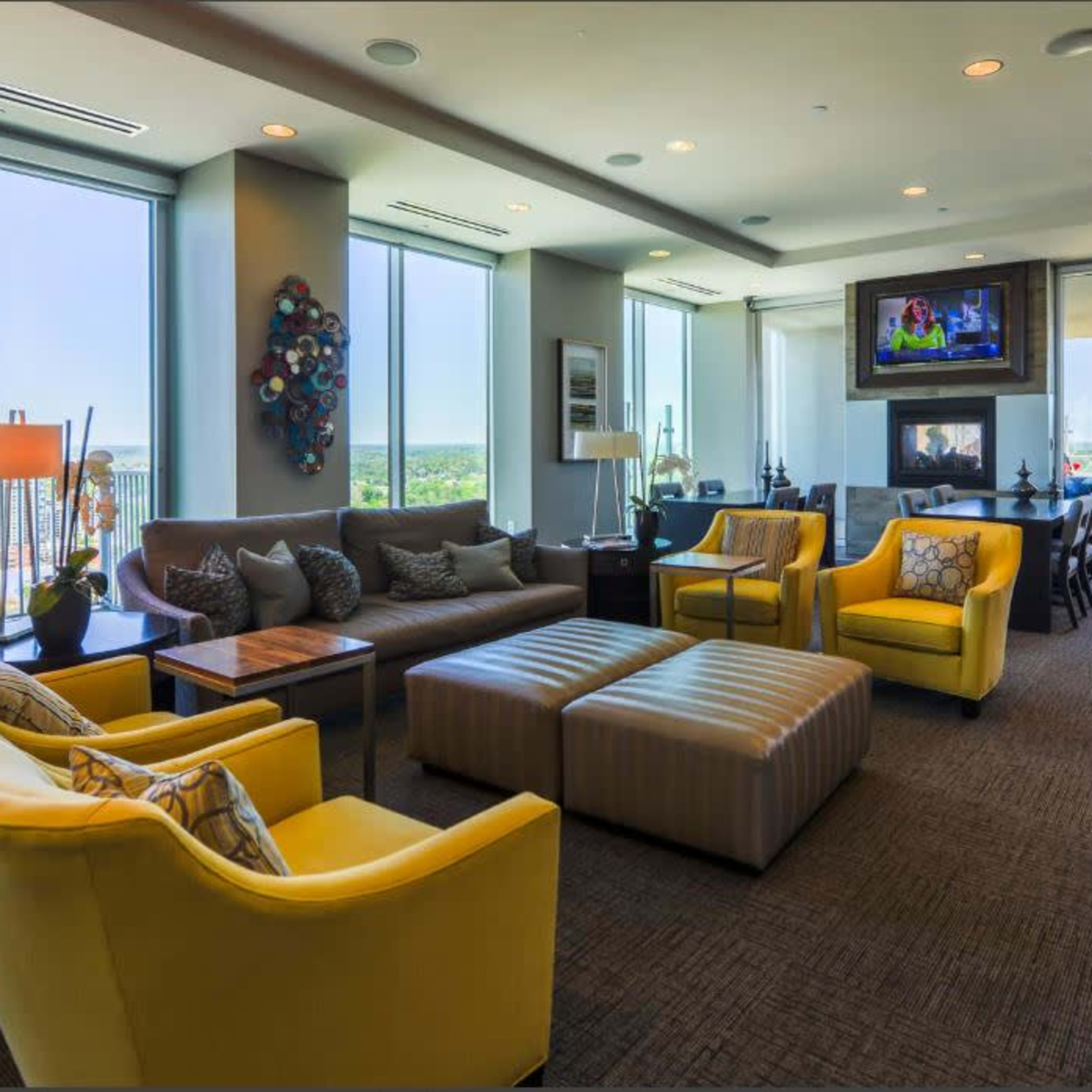 A modern living space with large windows, yellow armchairs, a sectional sofa, and a television mounted on the wall.