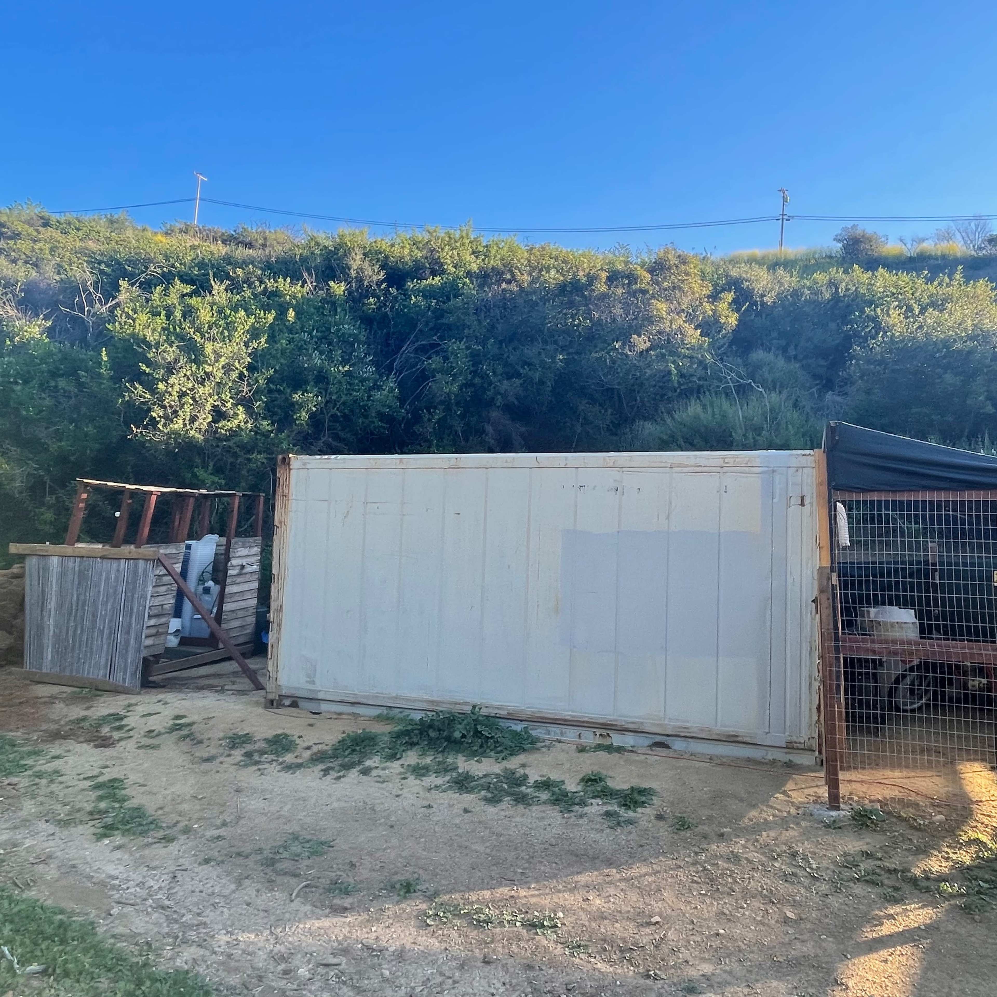 A white shipping container placed beside a fenced area on a grassy hillside.