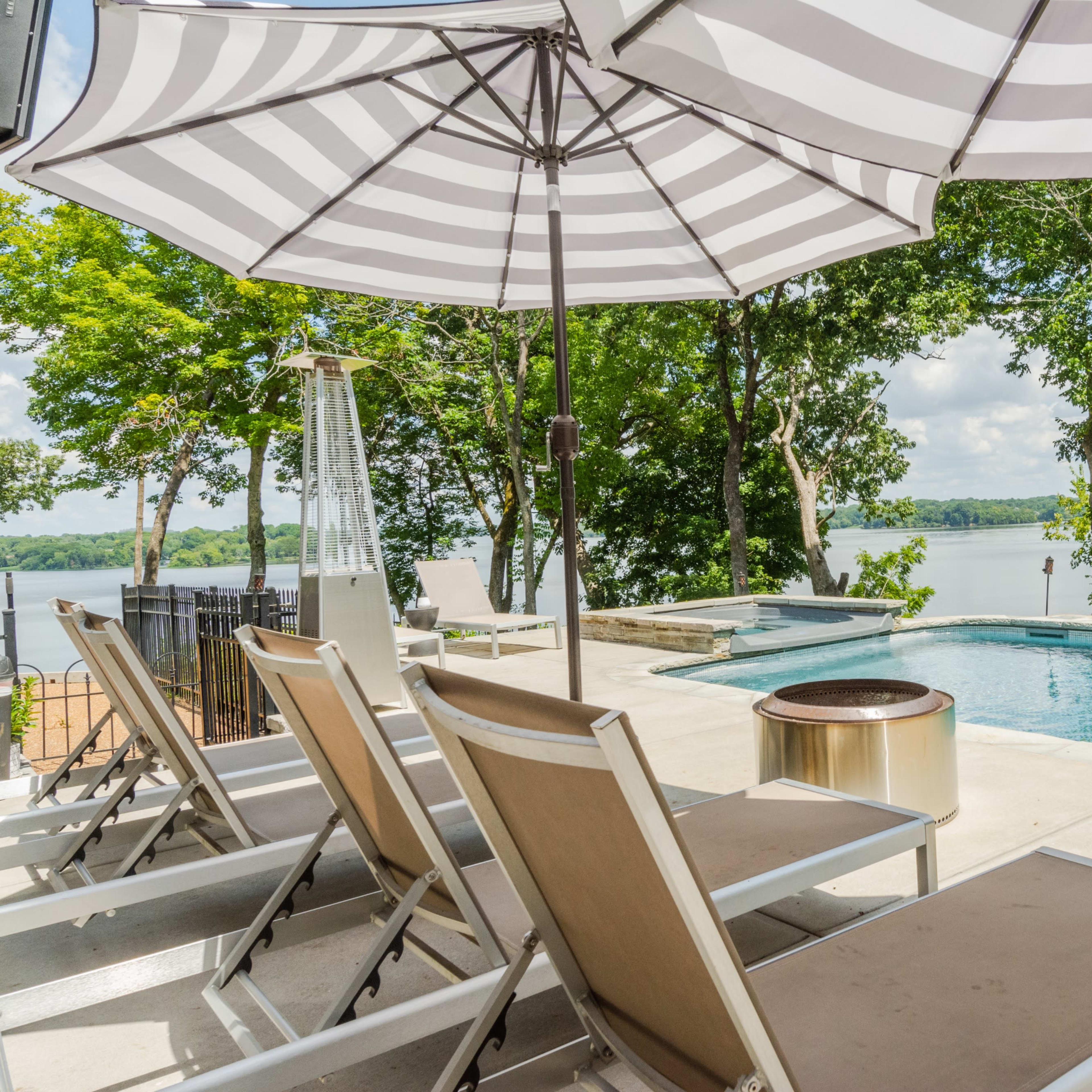 A pool area with lounge chairs and striped umbrellas overlooking a lake surrounded by trees.